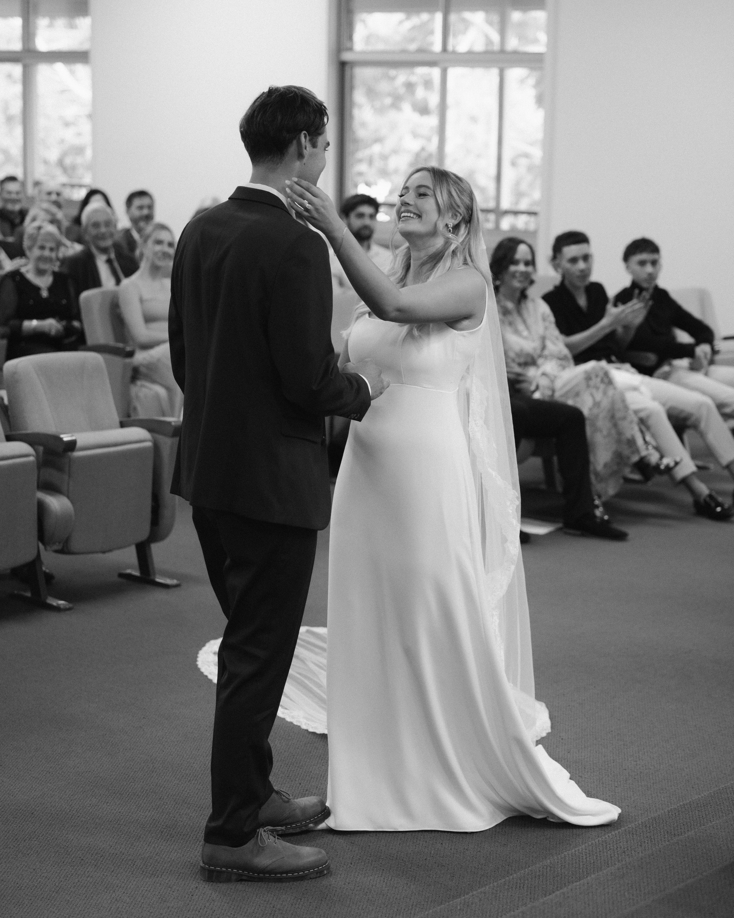 A bride and groom exchanging vows during a wedding ceremony indoors, with guests seated in the background.