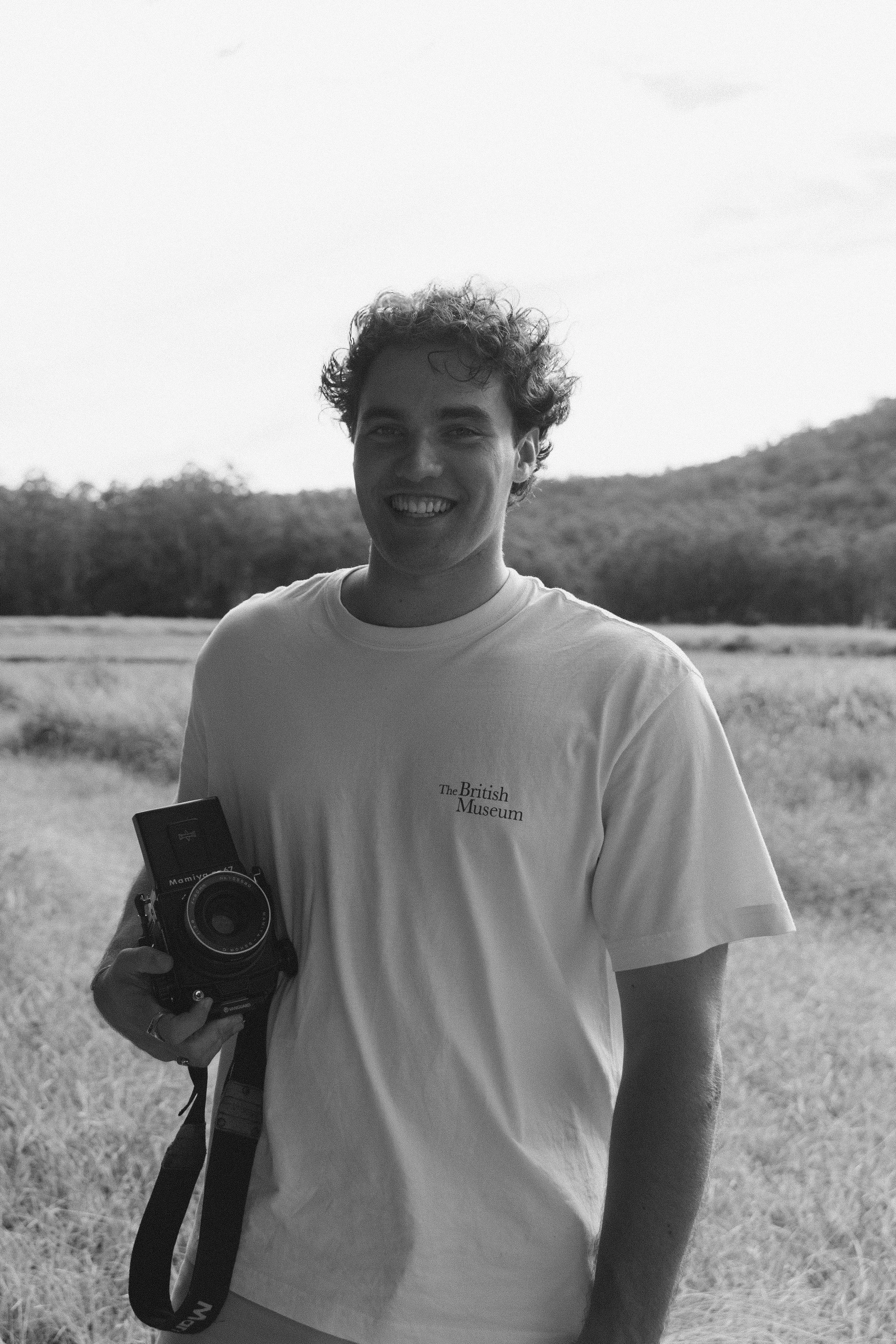 Young man outdoors in a field holding a camera, smiling, wearing a t-shirt with 'The British Museum' logo, black and white photograph.