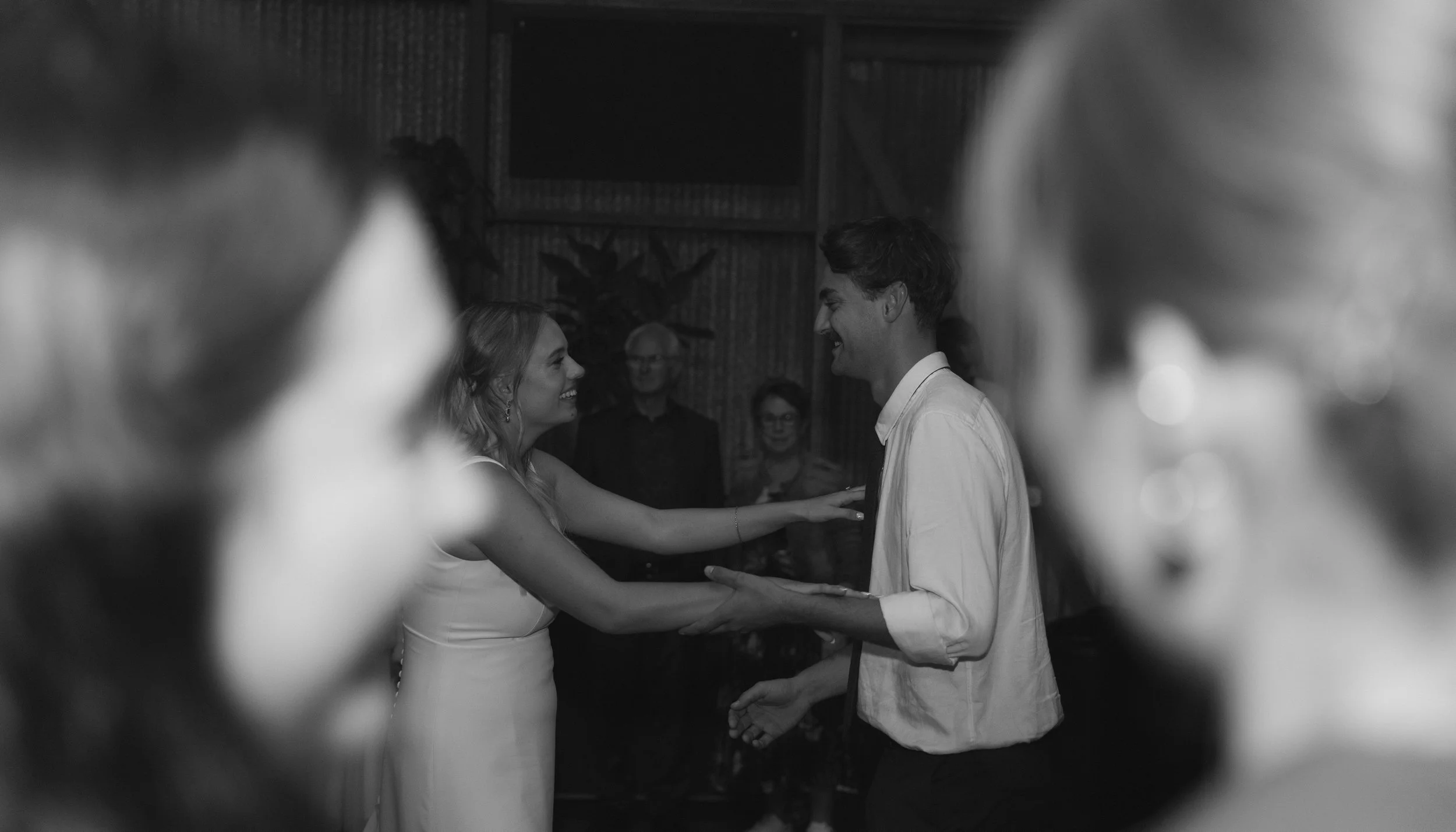 A black and white photo of a couple dancing, smiling at each other, with onlookers in the background. The scene appears to be at a celebration or party.