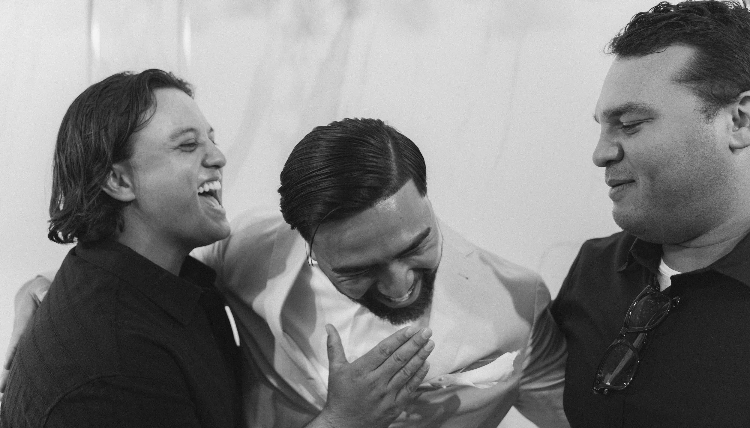 Three friends are laughing and smiling together, two men and one woman. They are close, with one person touching the other's chest, and appear to be sharing a joyful moment.