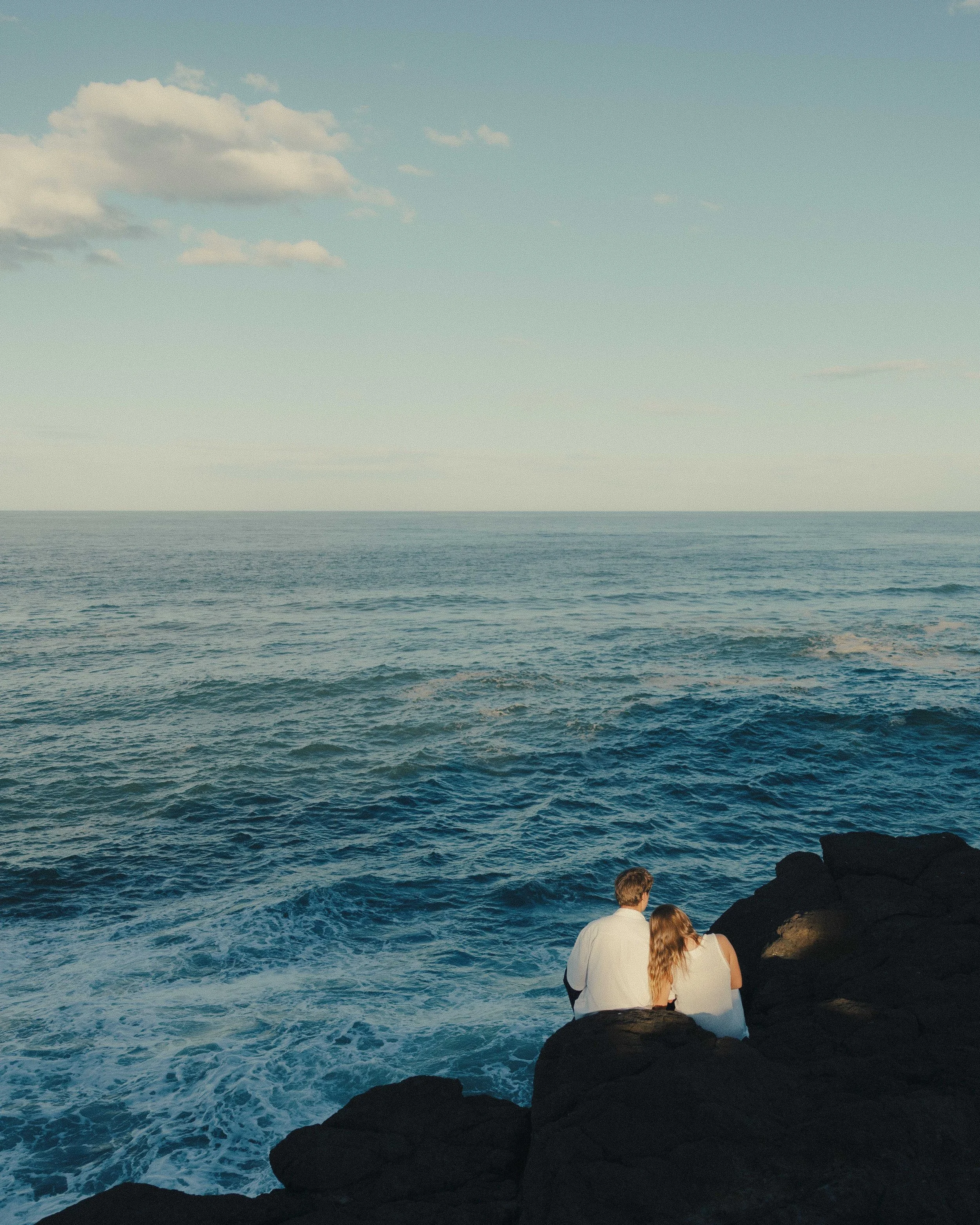 A couple sitting on rocks by the ocean, gazing at the water during sunset or sunrise.