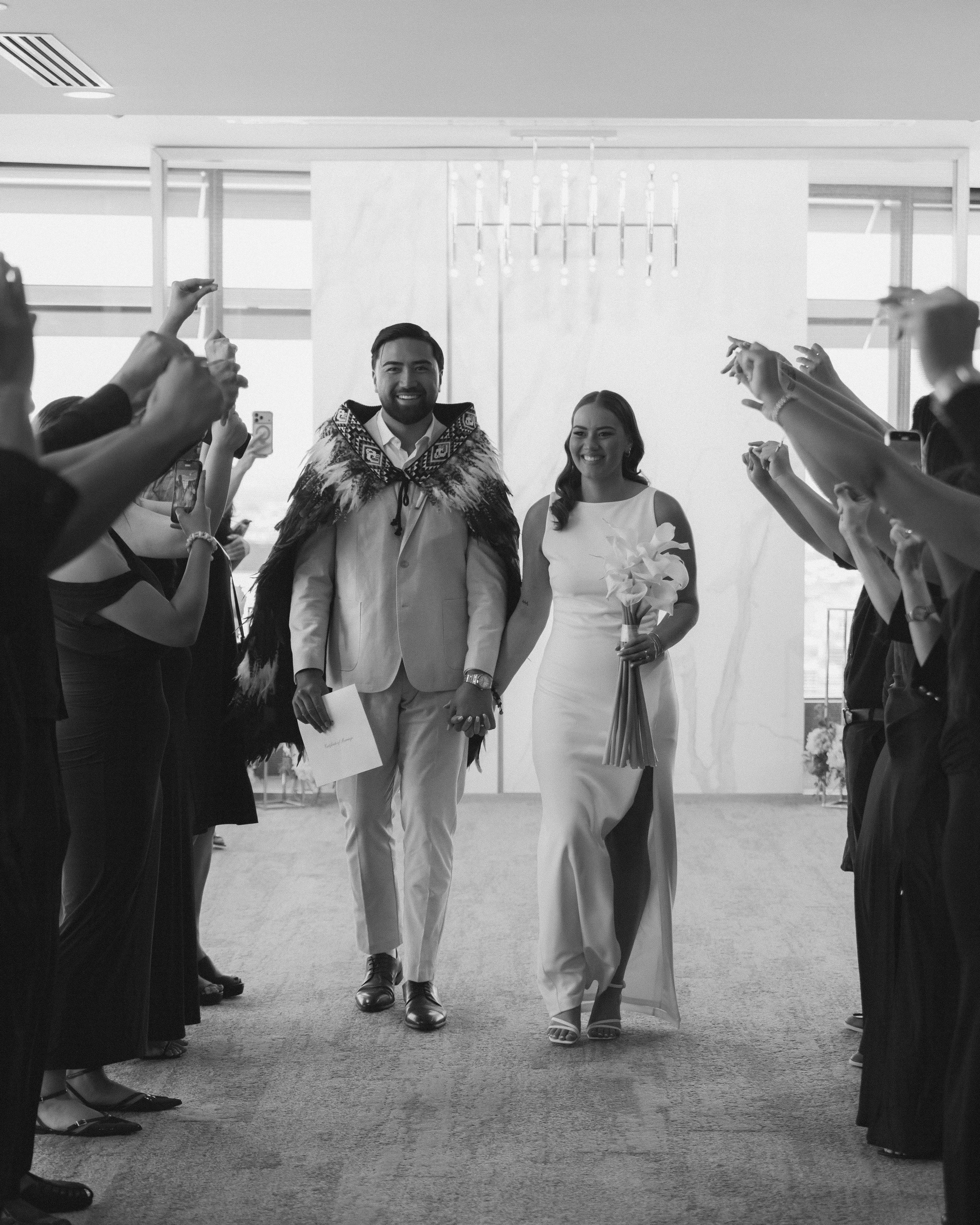A bride and groom holding hands, walking down the aisle during a wedding ceremony, surrounded by friends and family who are taking photos.