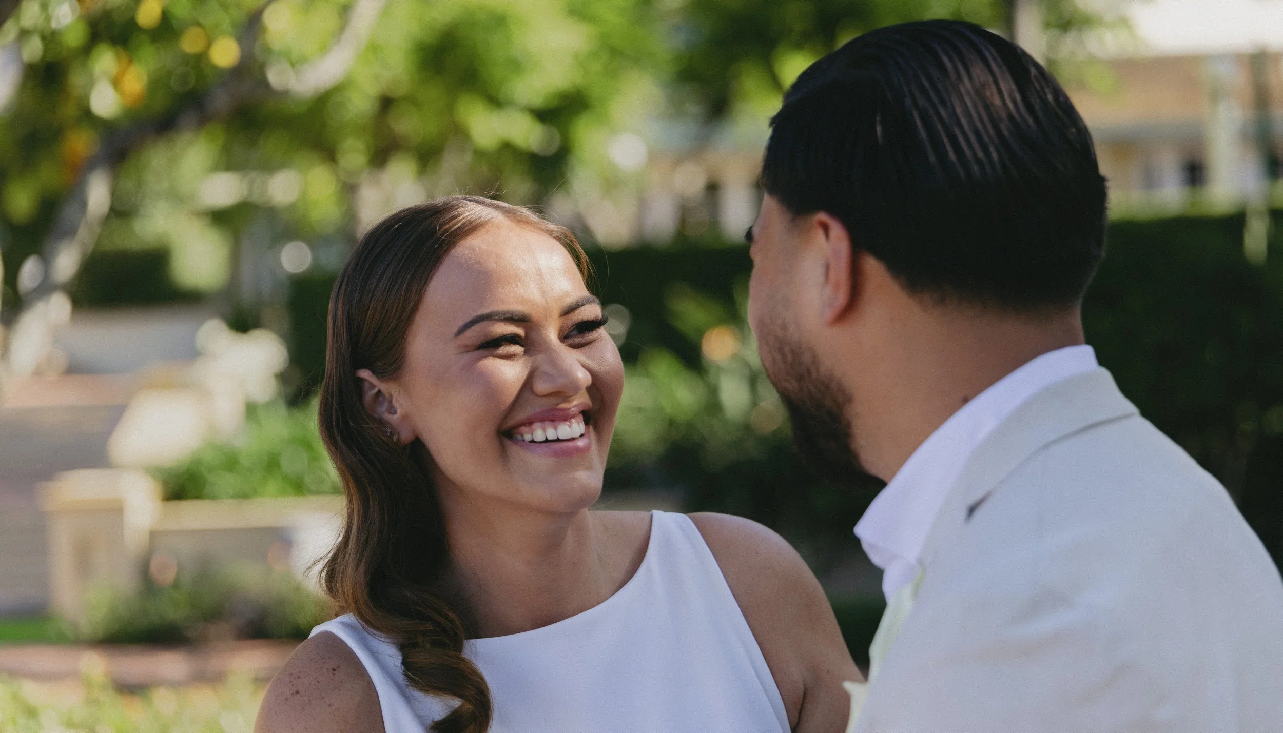 A woman and a man smiling and looking at each other outdoors in a park or garden setting.