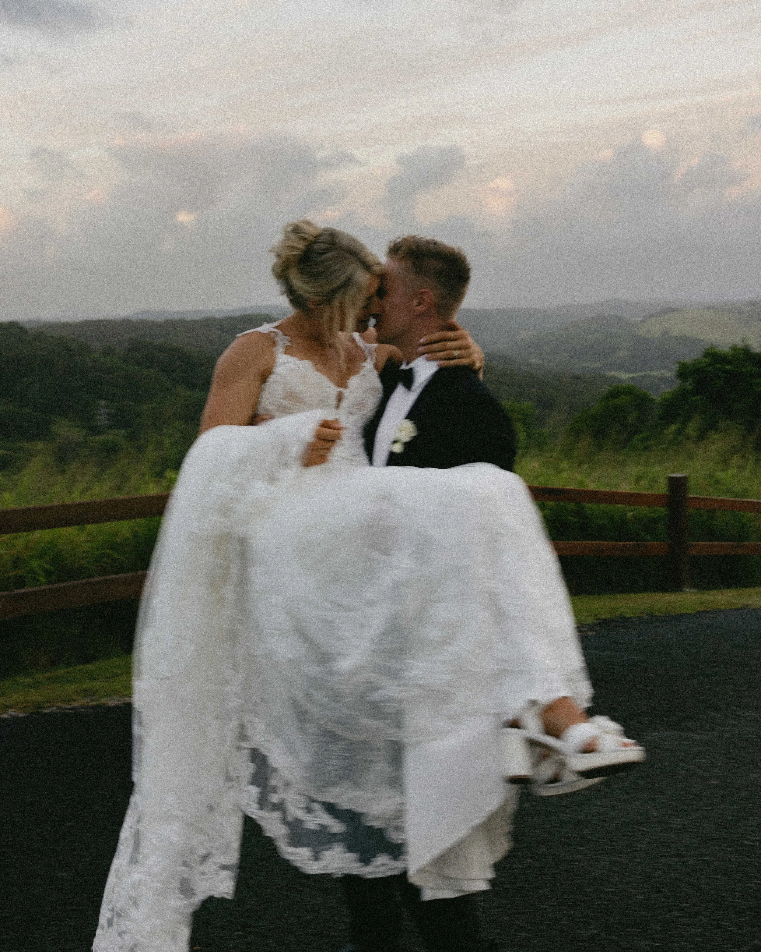 A bride and groom celebrating their wedding outdoors, with the groom lifting the bride and leaning in for a kiss, green hills and a cloudy sky in the background.