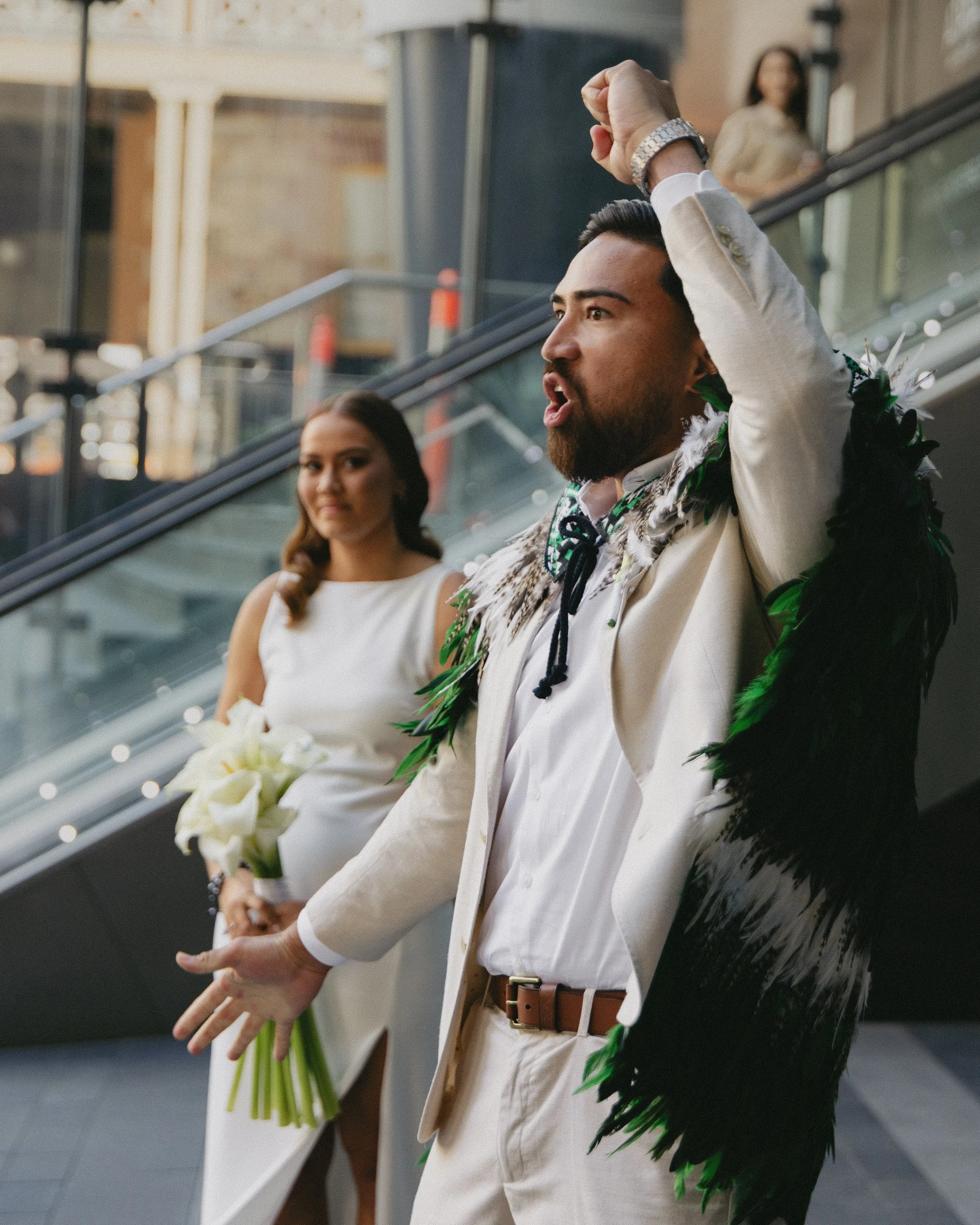 A man in a beige suit jacket and dress shirt plays the ukulele with his right arm raised, while a woman in a white dress holds a bouquet of calla lilies behind him. They are on an outdoor escalator in an urban setting, with a woman in yellow in the background on a higher level.