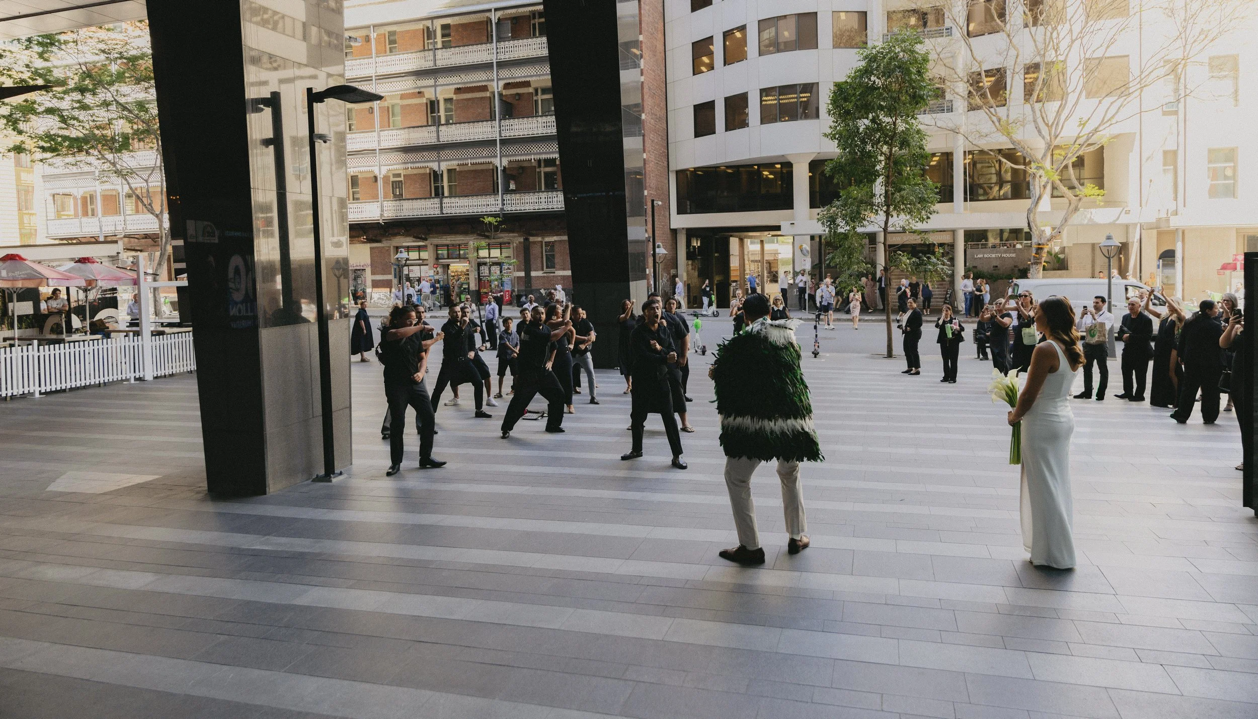 People gathered outside a building, some taking photos, a woman in a white dress holding flowers, and a man wearing a feathered costume, in an urban setting during daylight.
