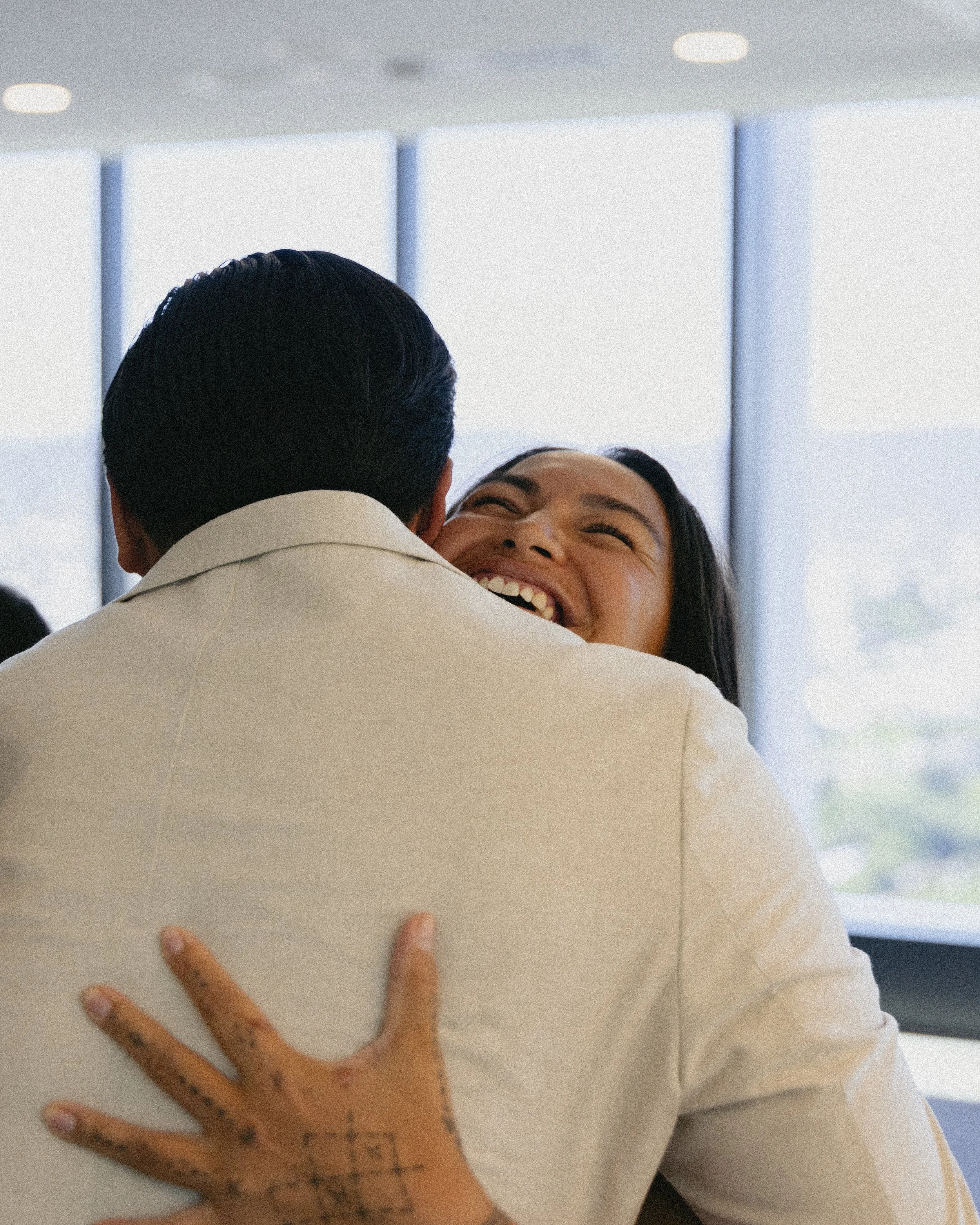 Two people hugging; one person's face is visible, smiling and laughing, while the back of the other's head and shoulder are visible.