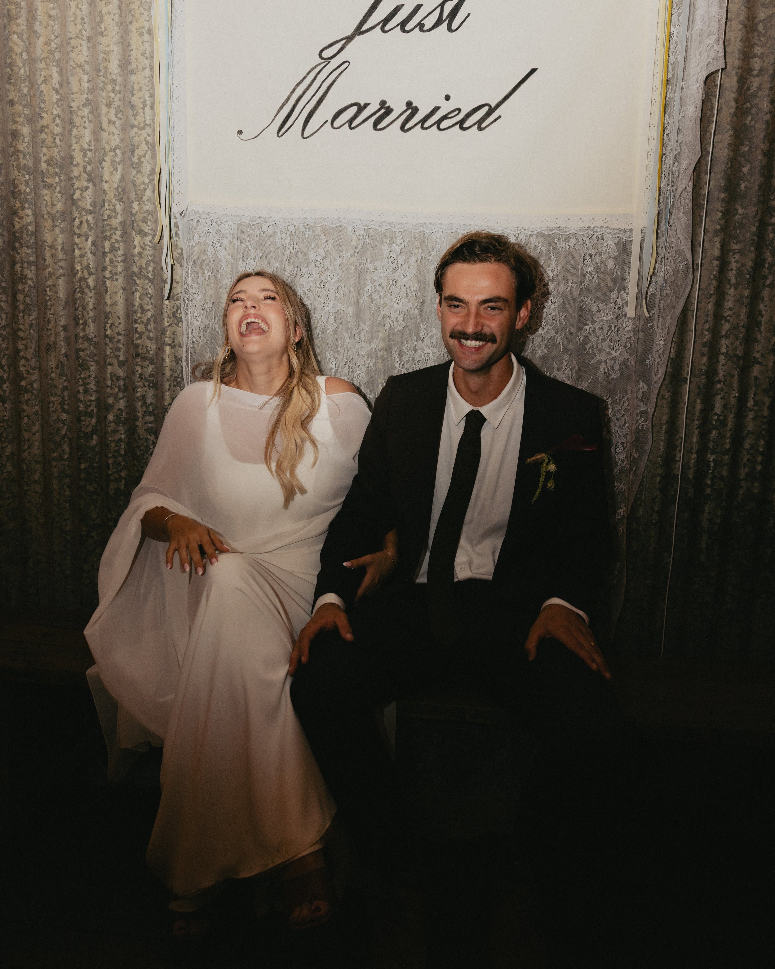 A bride and groom laughing and sitting on a bench at their wedding reception. The bride is wearing a white dress, and the groom is in a black suit with a tie. Behind them is a white fabric backdrop with a sign that says "just Married."