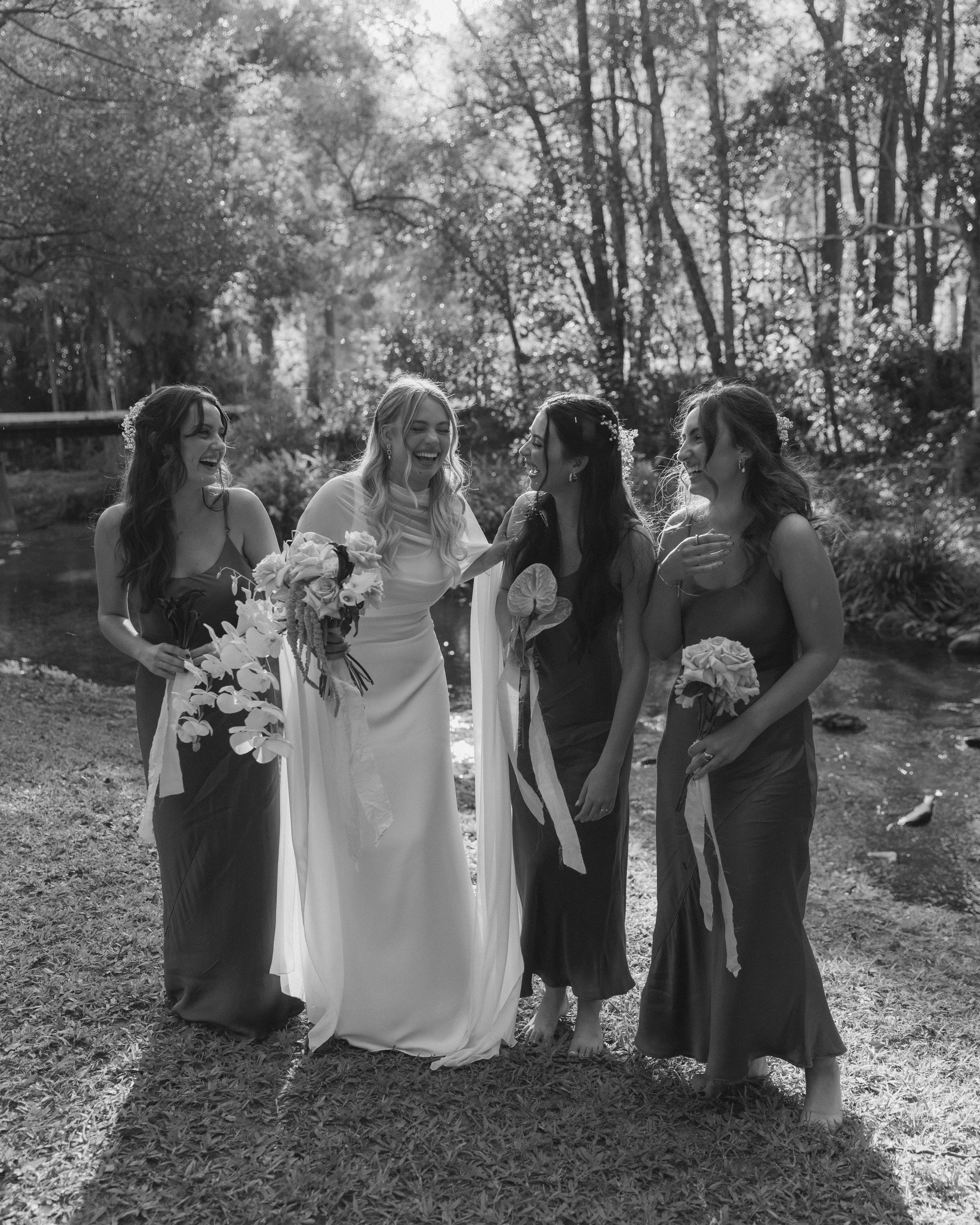 Group of five women, one in a wedding dress and four in dark dresses, smiling and laughing outdoors near a creek with trees in the background.
