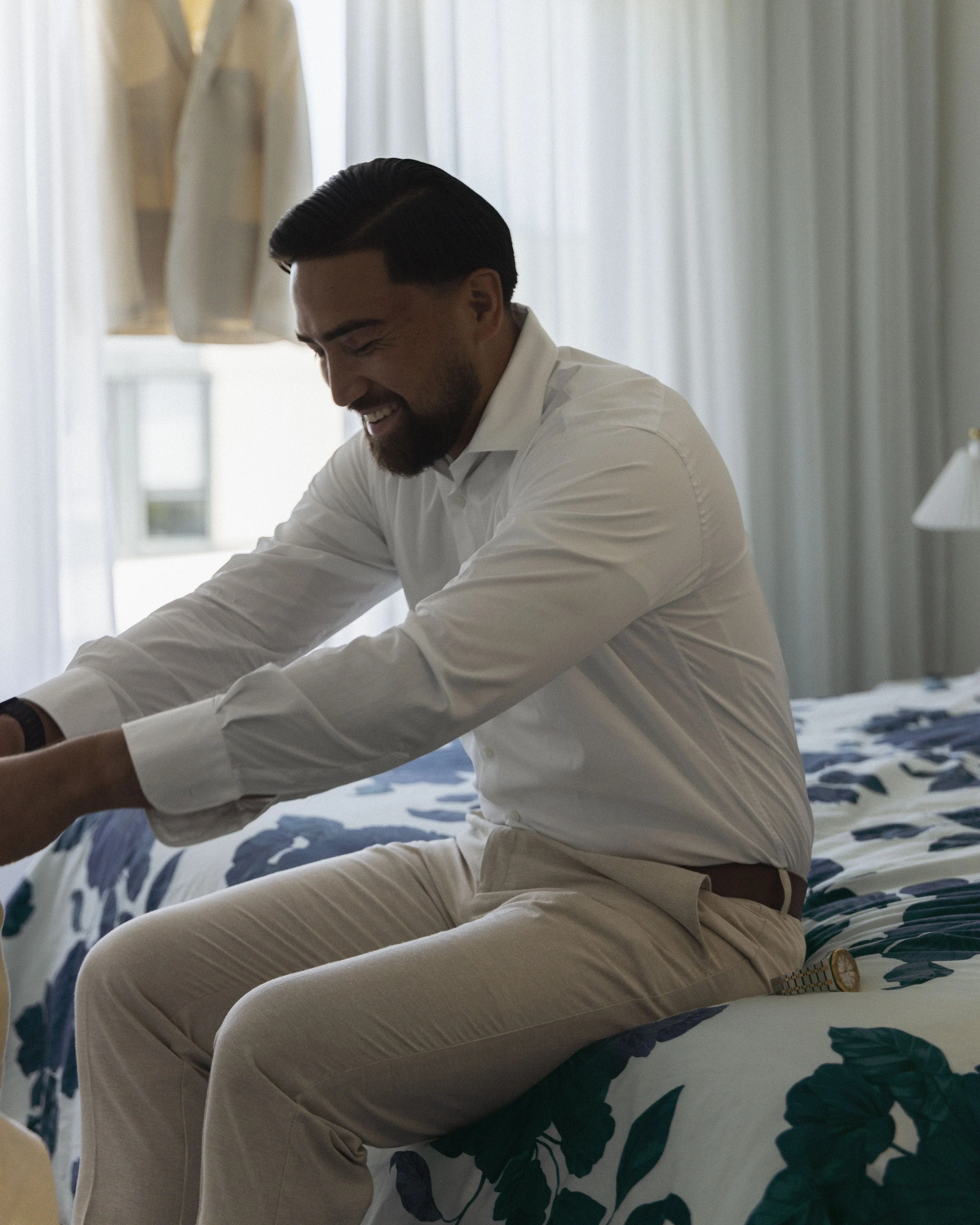 A man dressed in a white shirt and beige pants is sitting on a bed, smiling while getting dressed or undressed.