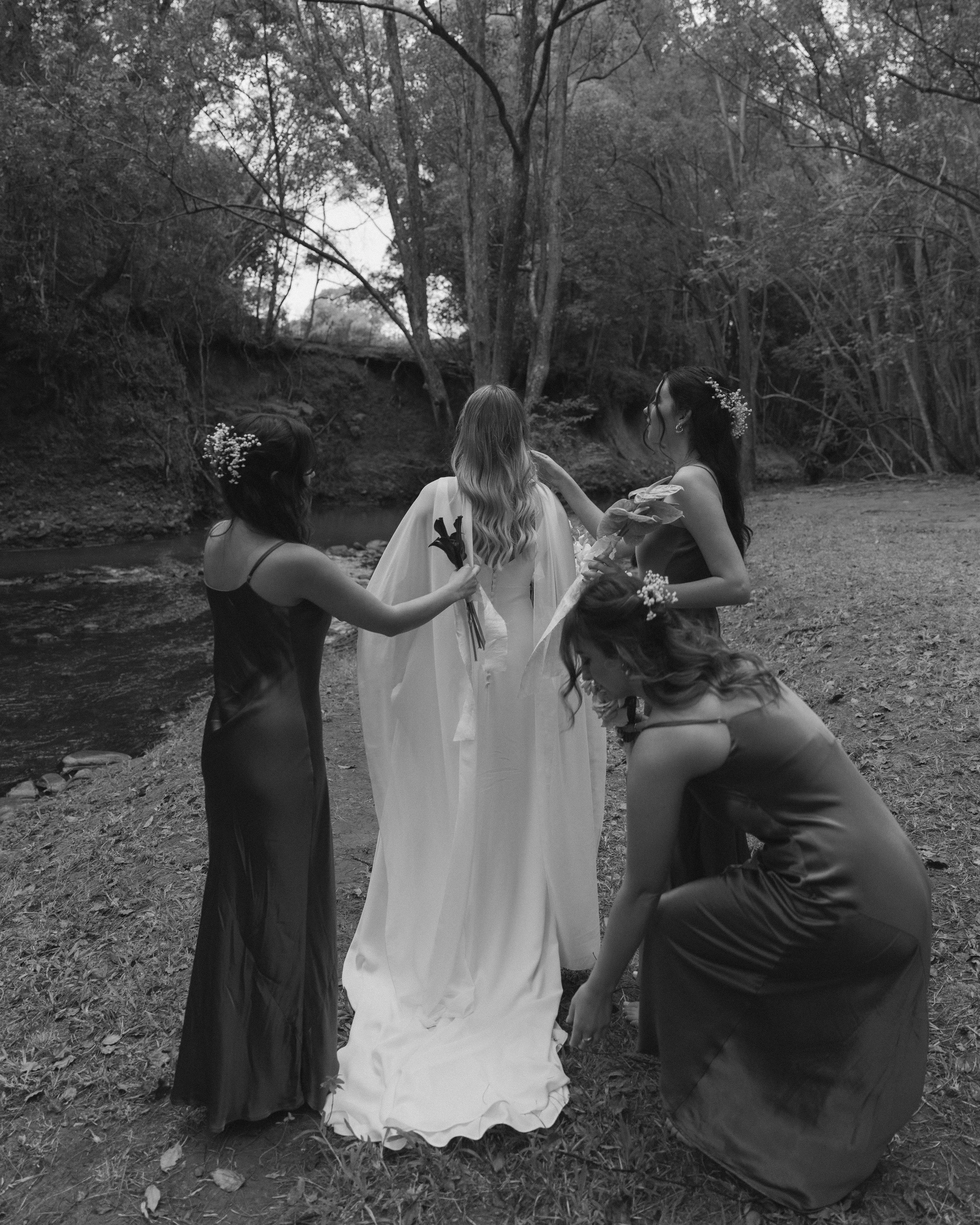 A group of four women, one in a white dress, in a wooded outdoor setting, with one woman kneeling and the others standing around her.