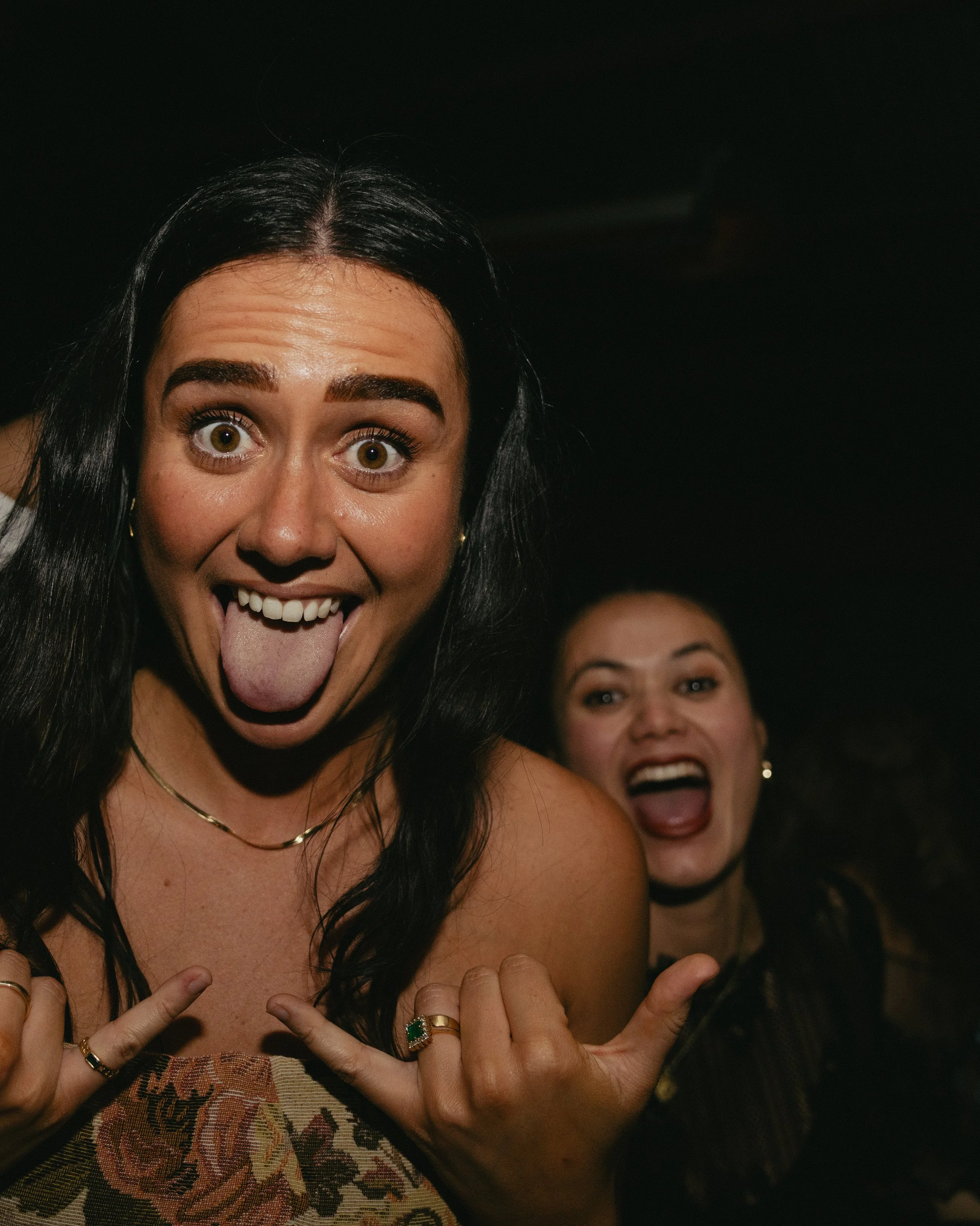 Two women smiling and making playful gestures with their tongues out, close-up shot, dark background.