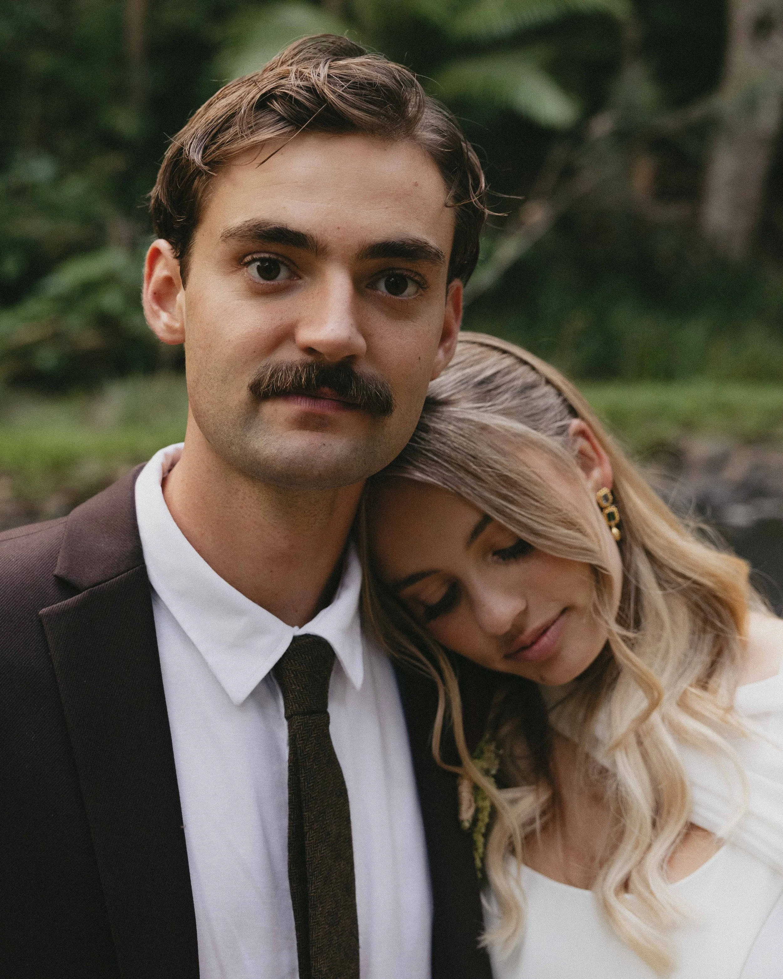 A man with a mustache wearing a suit and tie, and a woman with blonde hair in a white dress, resting her head on his shoulder outdoors with greenery in the background.