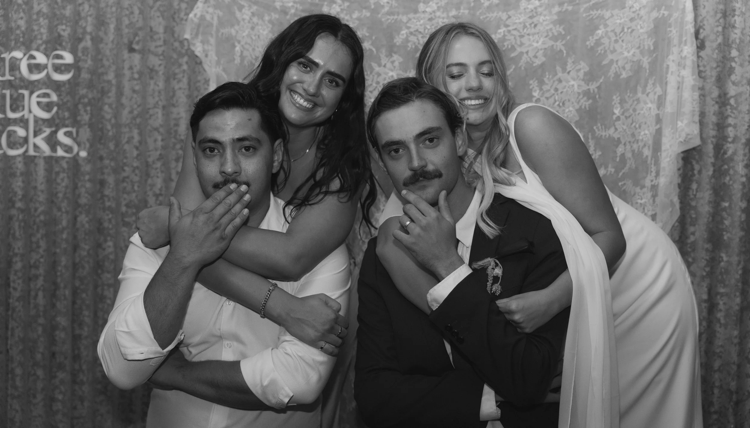 Four people posing closely together in front of a decorative backdrop; two women and two men, with the women hugging the men from behind.