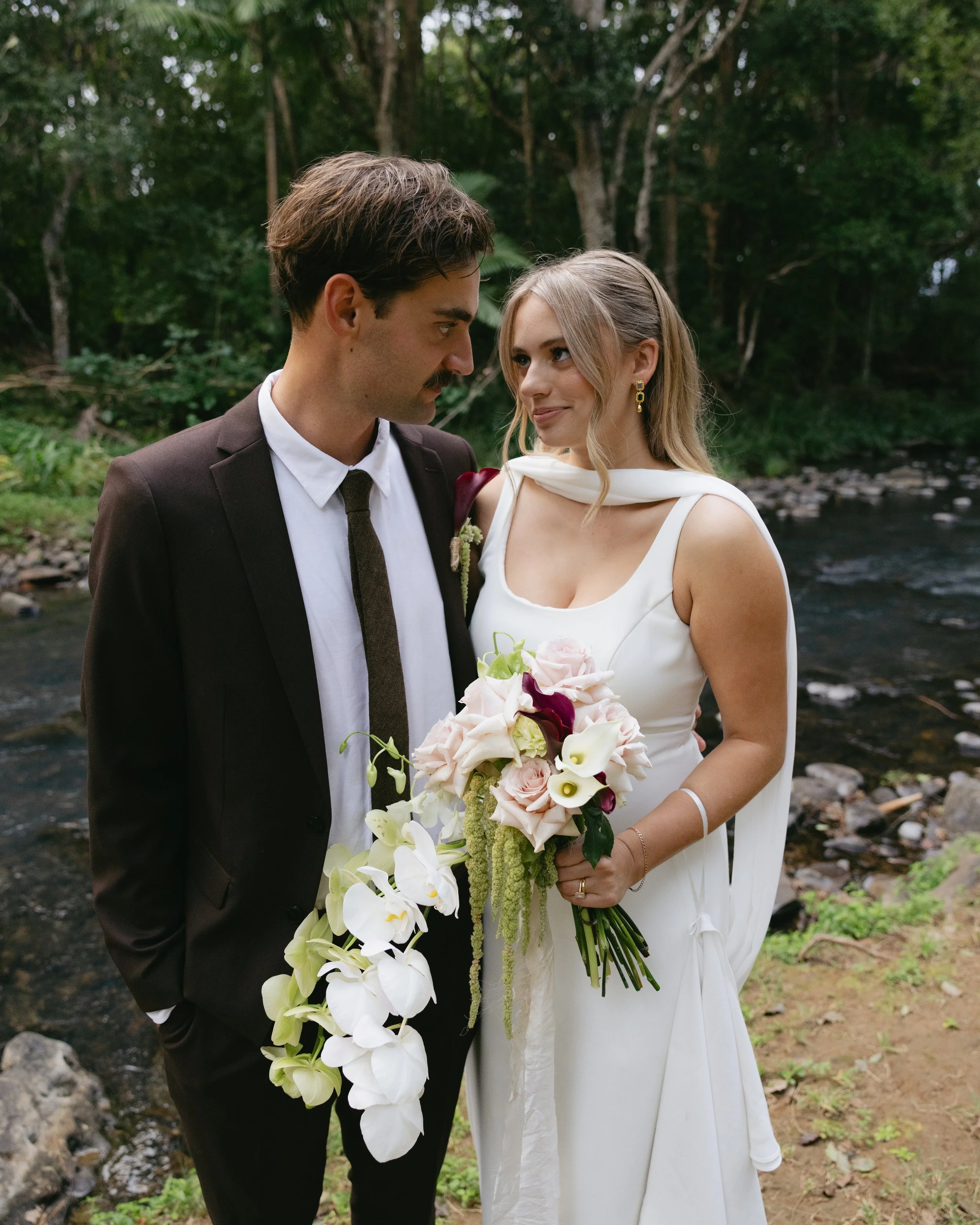 Bride and groom in wedding attire standing near a river in a wooded area, holding a bouquet of flowers.