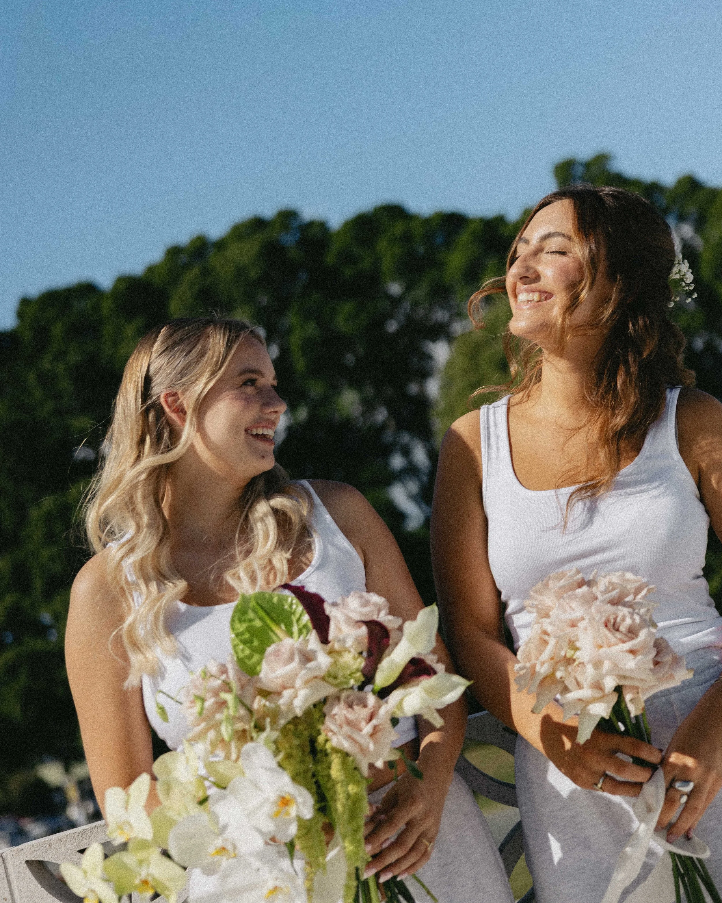 Two women smiling and holding flower bouquets outdoors on a sunny day, with trees in the background.