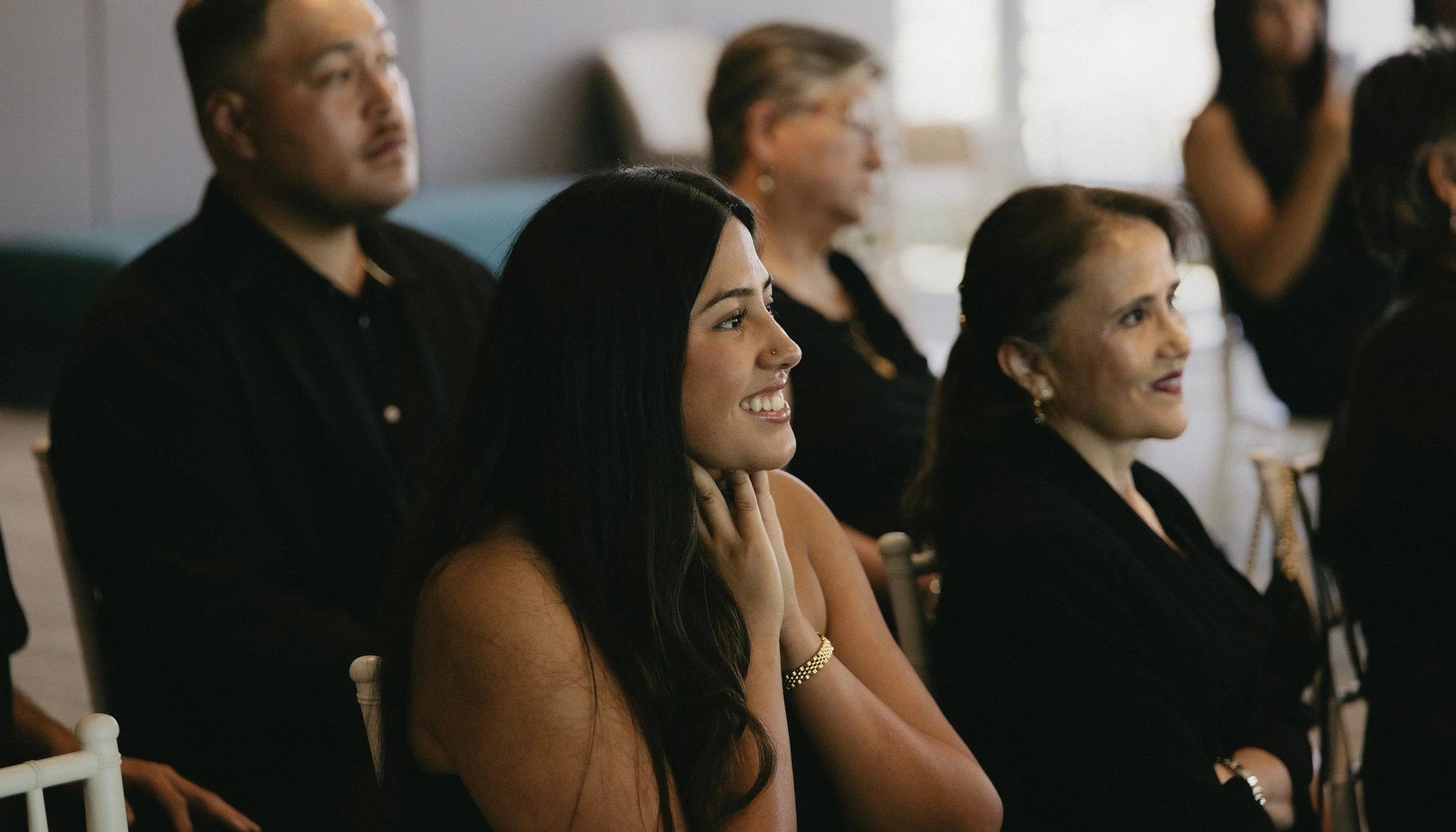 Group of well-dressed people seated and attentively listening at an indoor event or seminar, with natural light coming through window blinds in the background.
