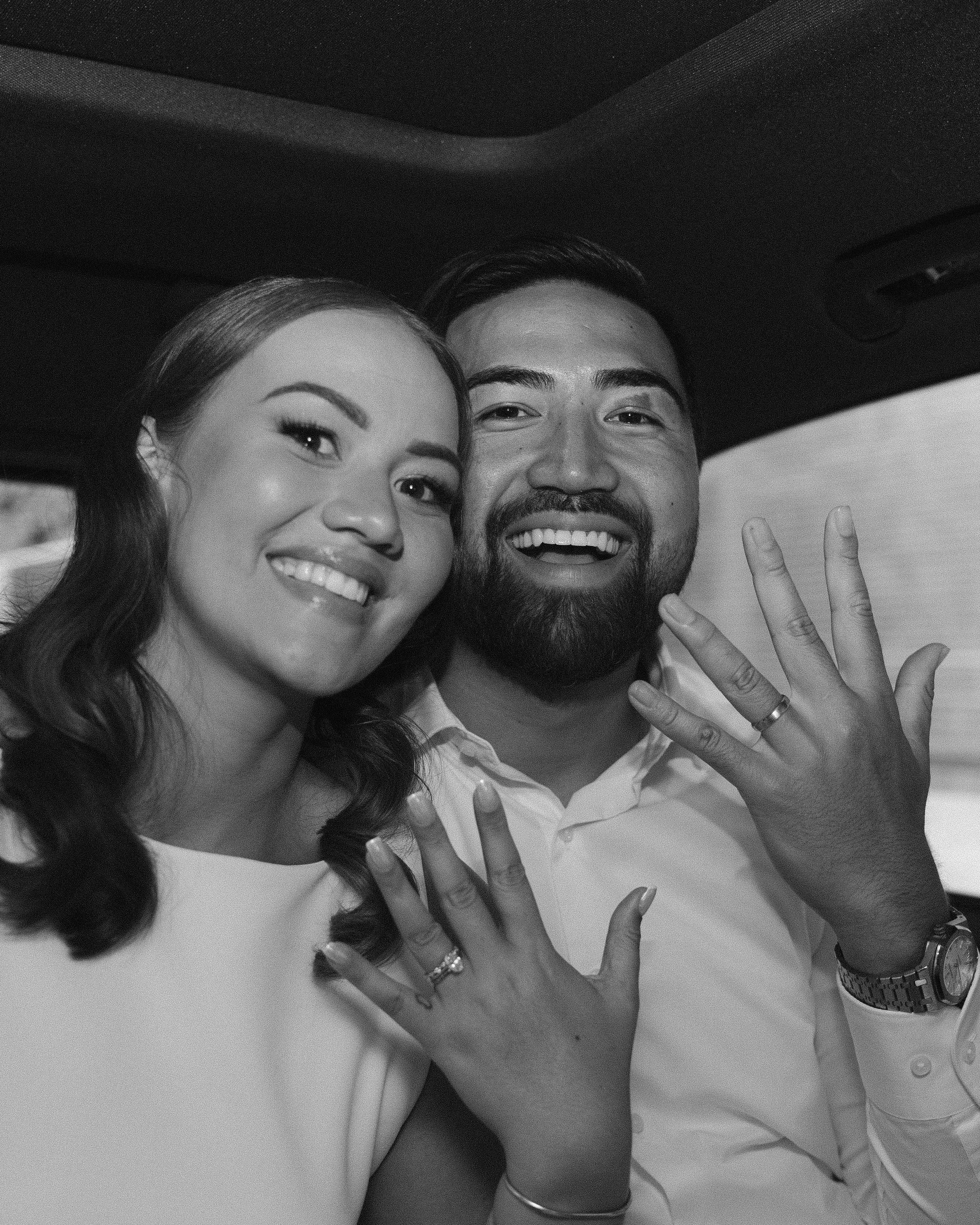 A smiling couple, the woman with wavy hair and the man with a beard, showing off rings on their hands, inside a vehicle.