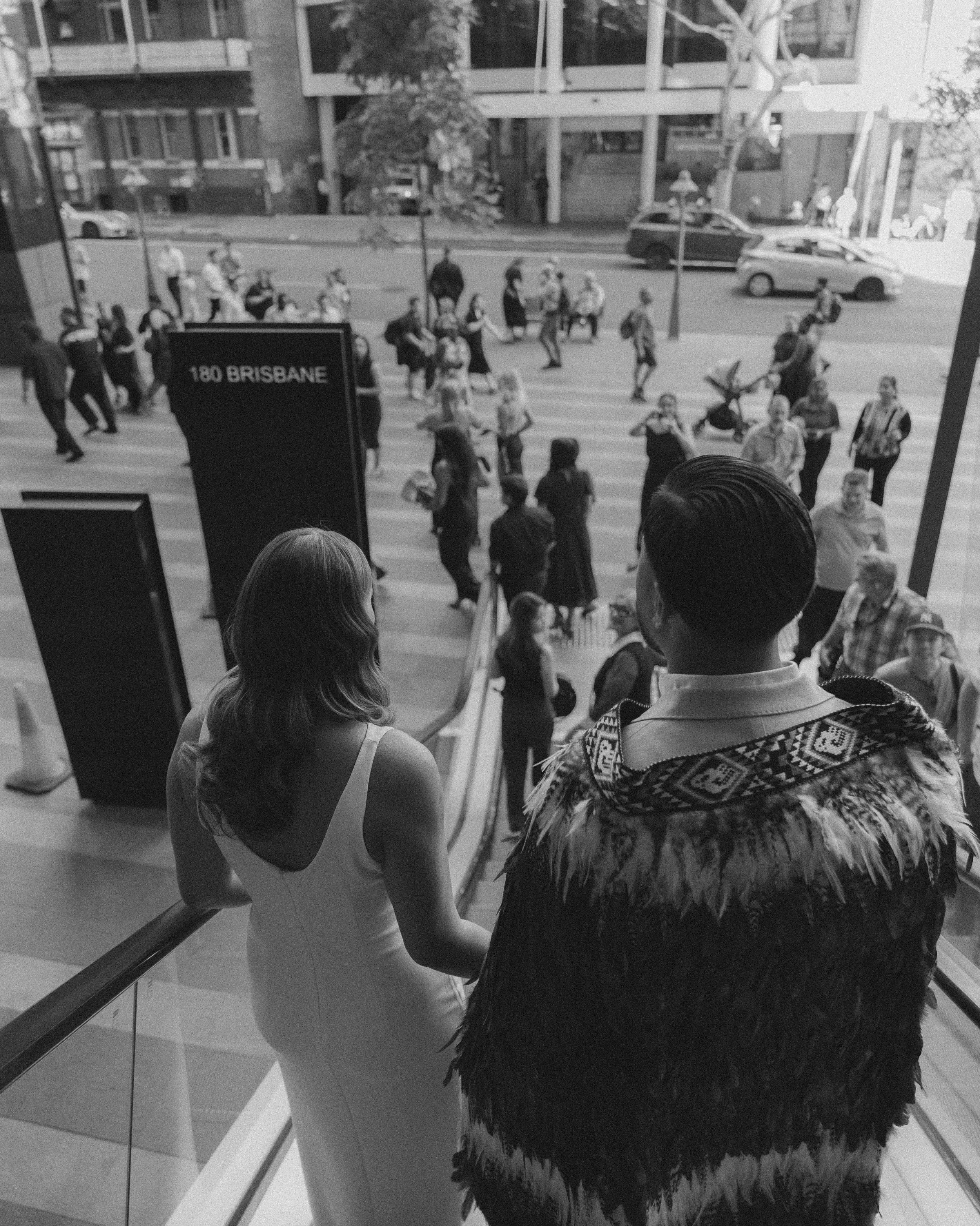 Two people look out from an indoor balcony at a busy city street with pedestrians and parked cars, with a sign displaying '180 BRISBANE'.
