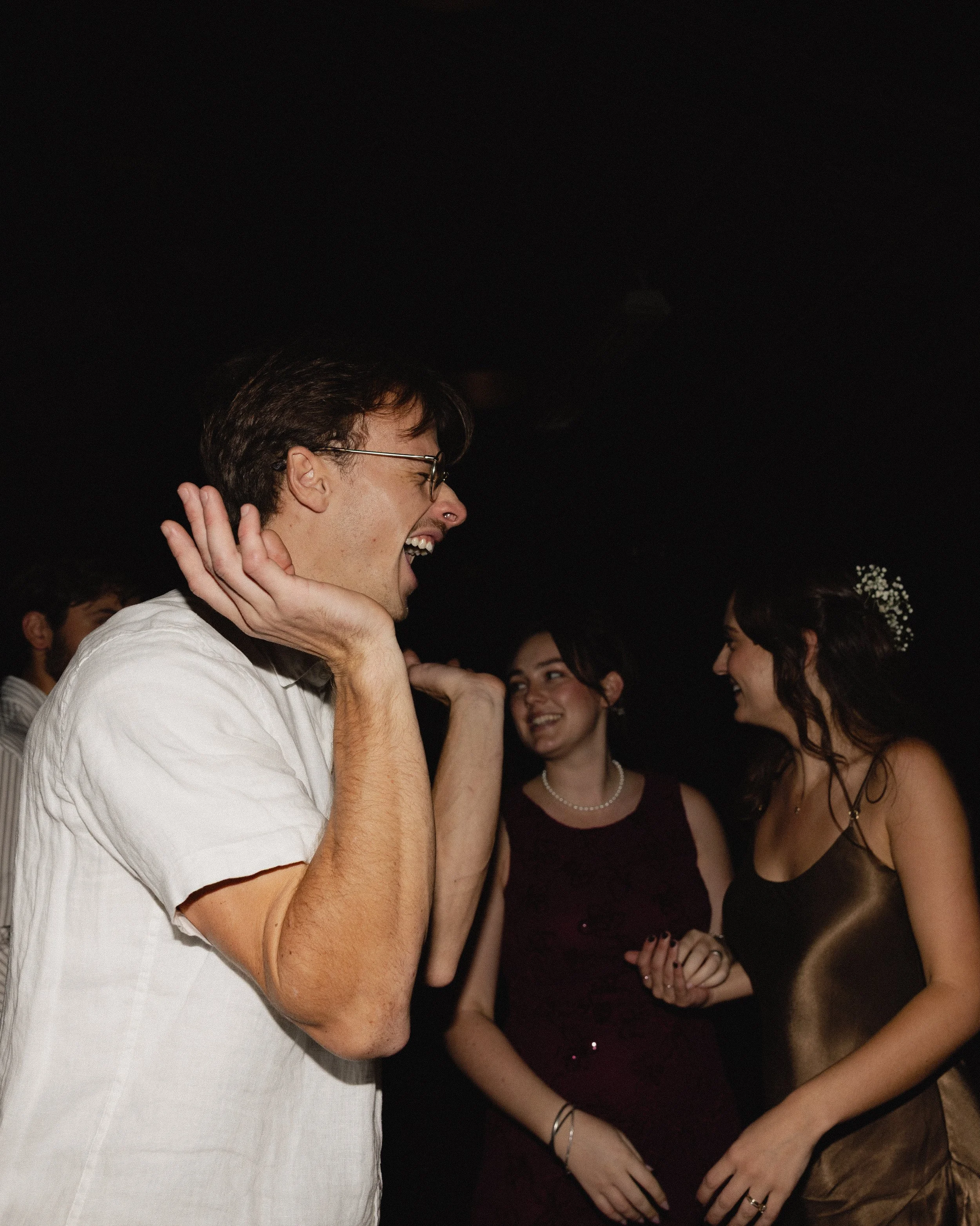 People laughing and talking at a celebration or party, with two women smiling and a man covering his ears, in a dimly lit room.