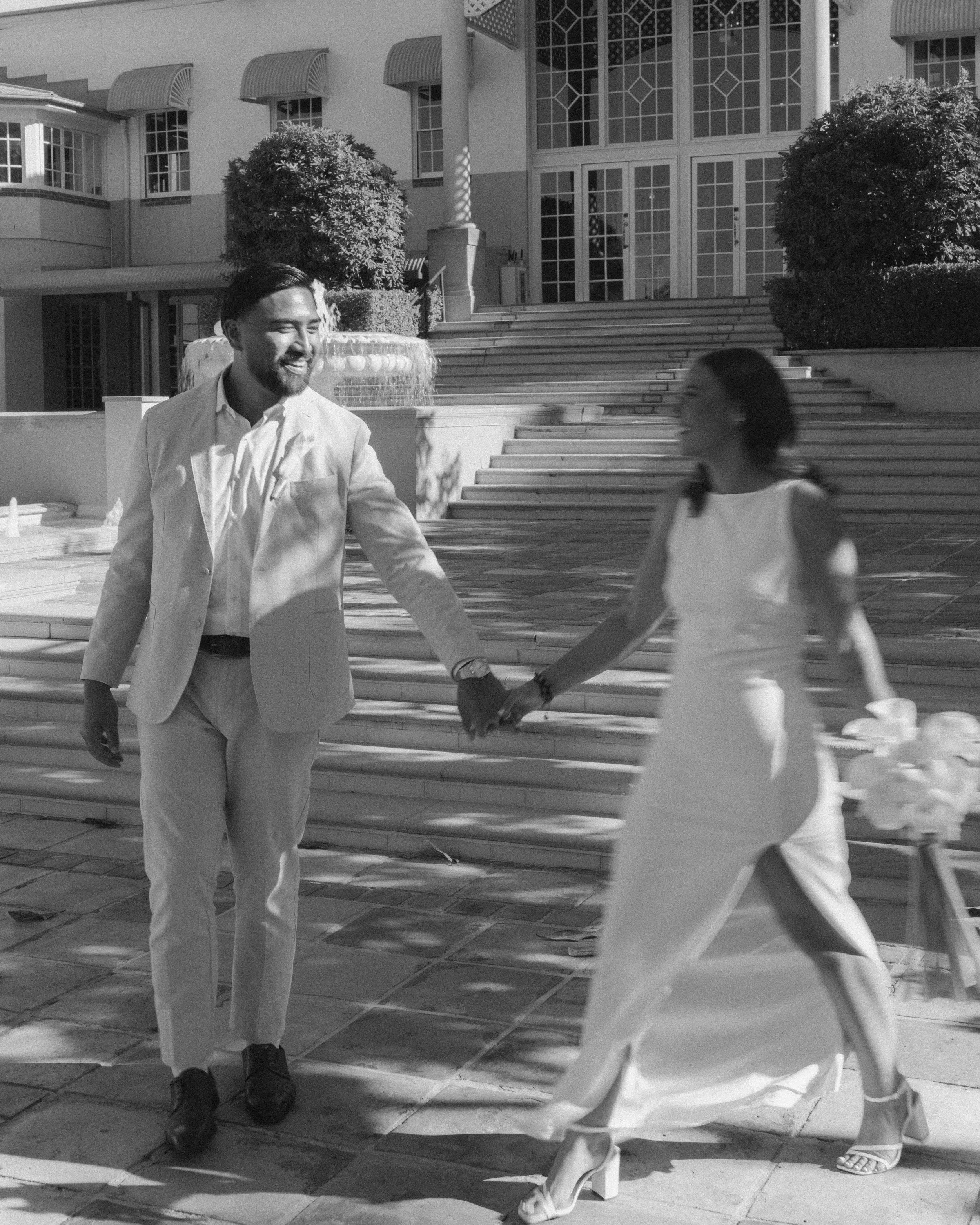 A man and woman are holding hands and walking outside in front of a building with stairs and windows, smiling at each other.
