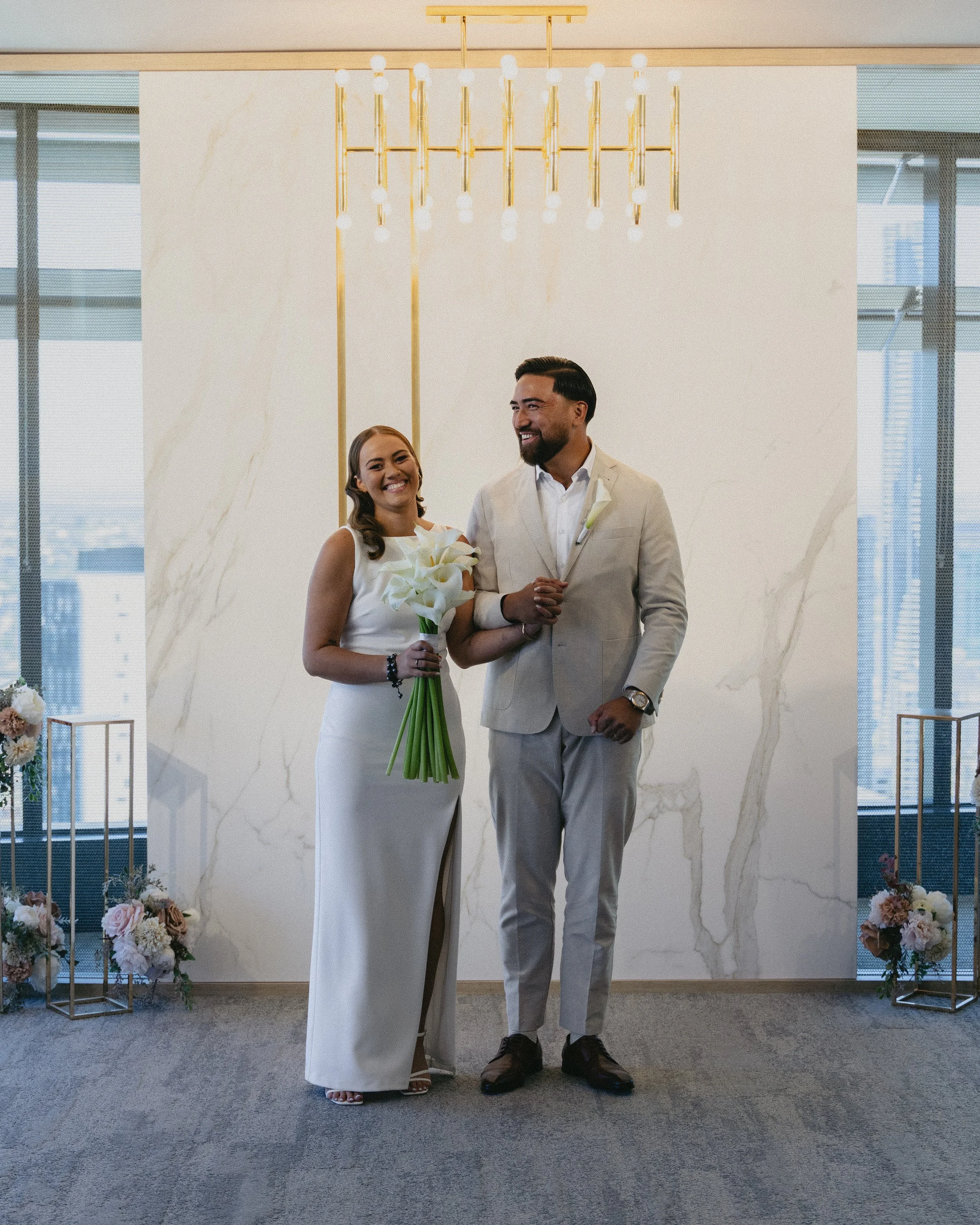 A happy couple, dressed in wedding attire, standing indoors with city skyscrapers visible through large windows behind them. The bride is holding a bouquet of white flowers and the groom is wearing a light suit. They are smiling and standing close together in a modern, elegant setting with floral decorations.