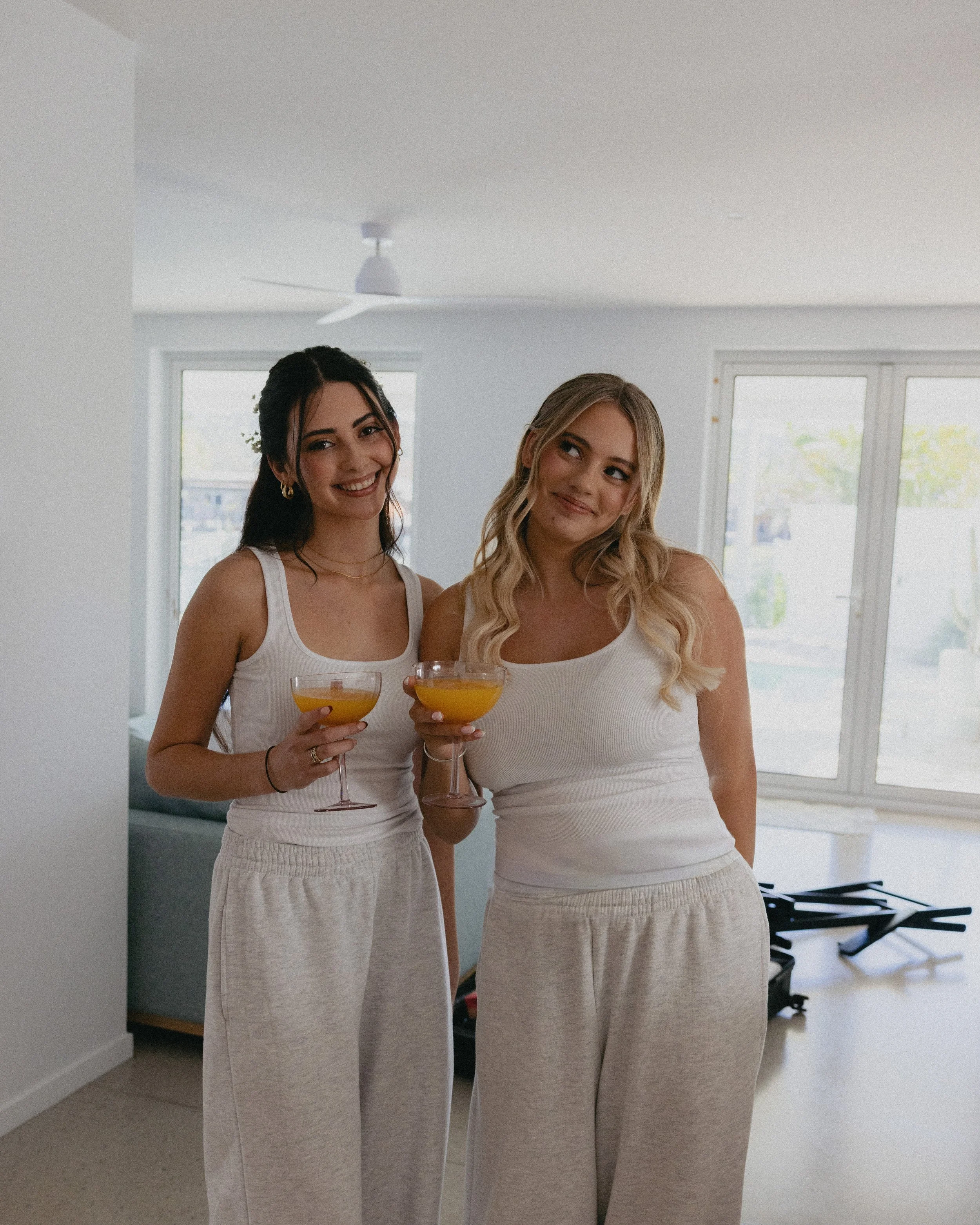 Two women standing indoors, holding drinks, smiling, wearing white tank tops and light-colored pants, with large windows and a ceiling fan in the background.