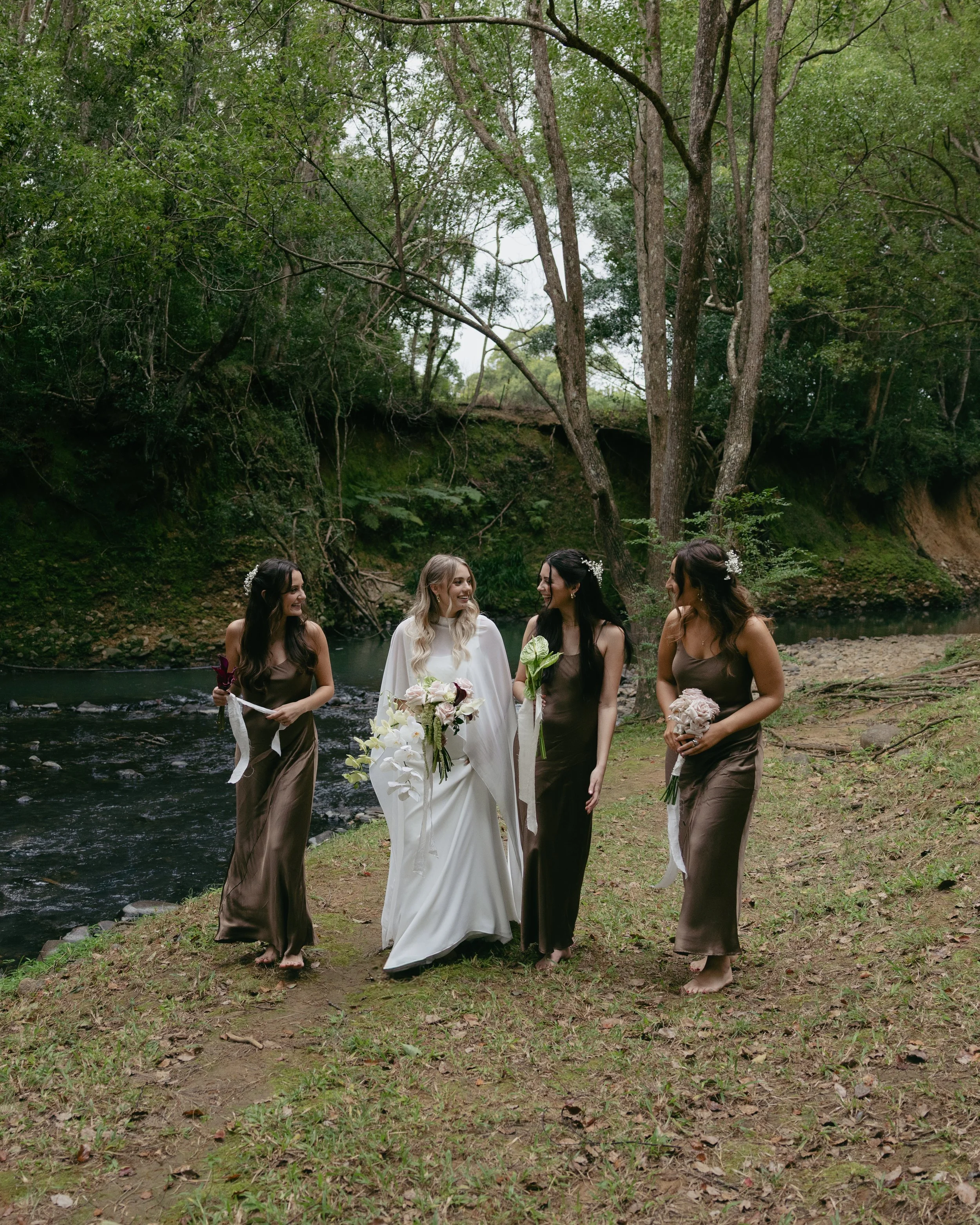 A bride and three bridesmaids walking barefoot along a riverbank in a forest.