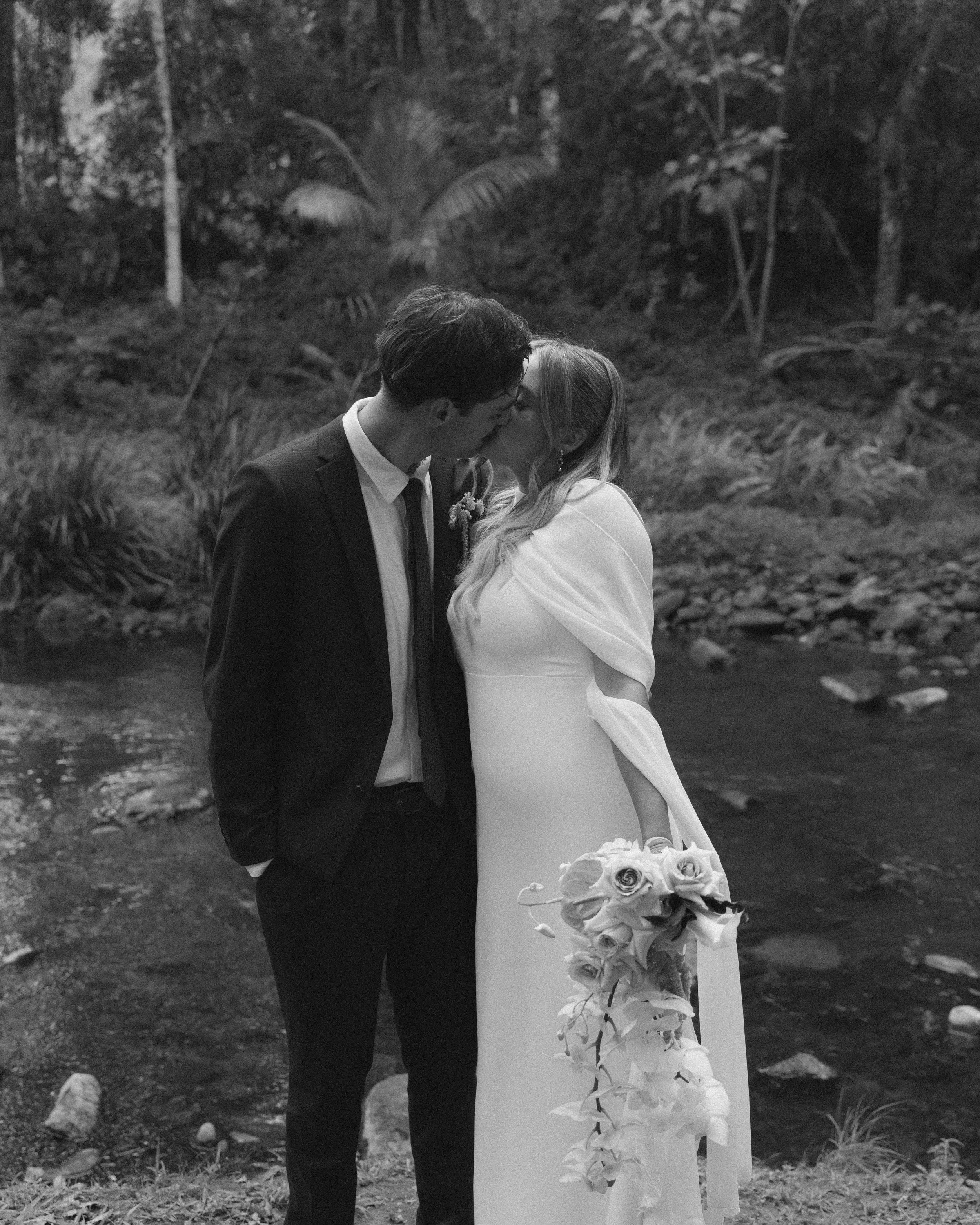 A black and white photo of a couple kissing outdoors by a stream, with the woman holding a bouquet of flowers.