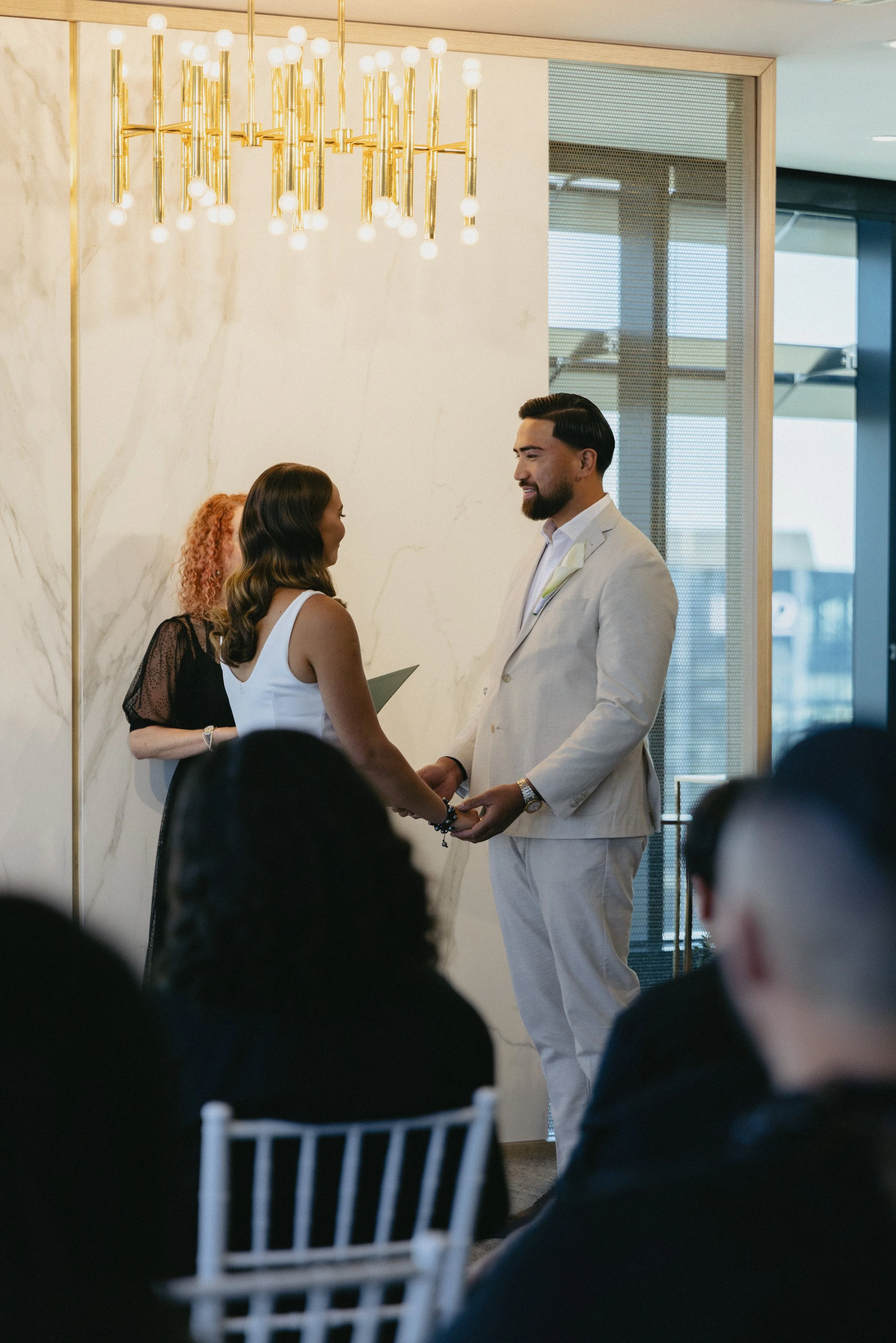 A couple gets married, holding hands and smiling at each other during their wedding ceremony in a decorated indoor space with a chandelier and guests seated in front.