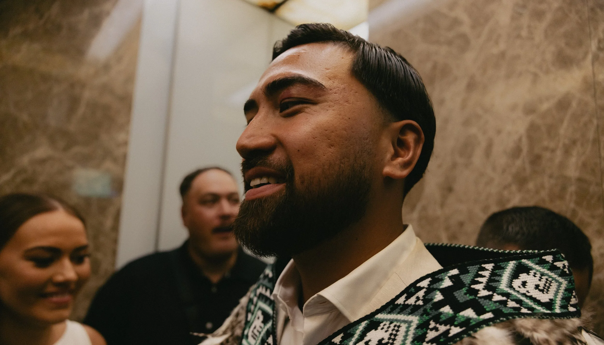 Close-up of a smiling man with a beard, wearing traditional clothing, at a social gathering indoors with a marble wall in background. Two other people, a woman and a man, are partially visible and blurred behind him.