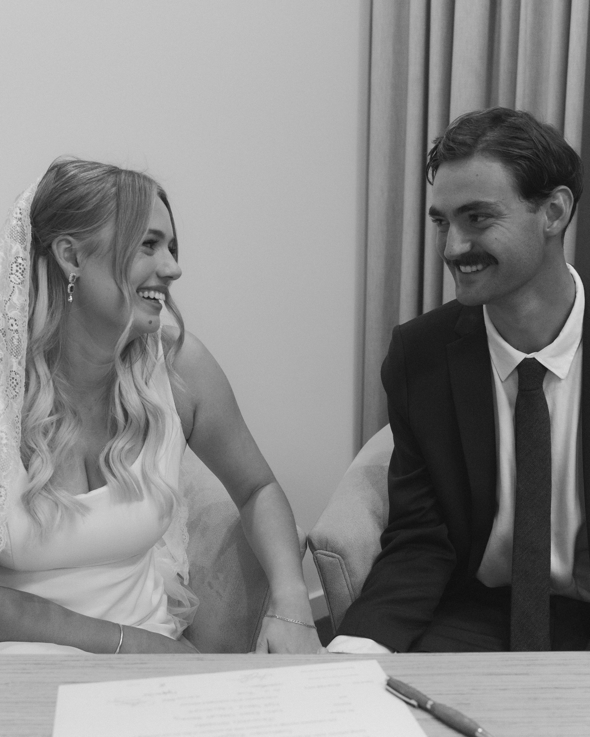 A bride and groom smiling at each other during their wedding ceremony, holding hands and sitting at a table.