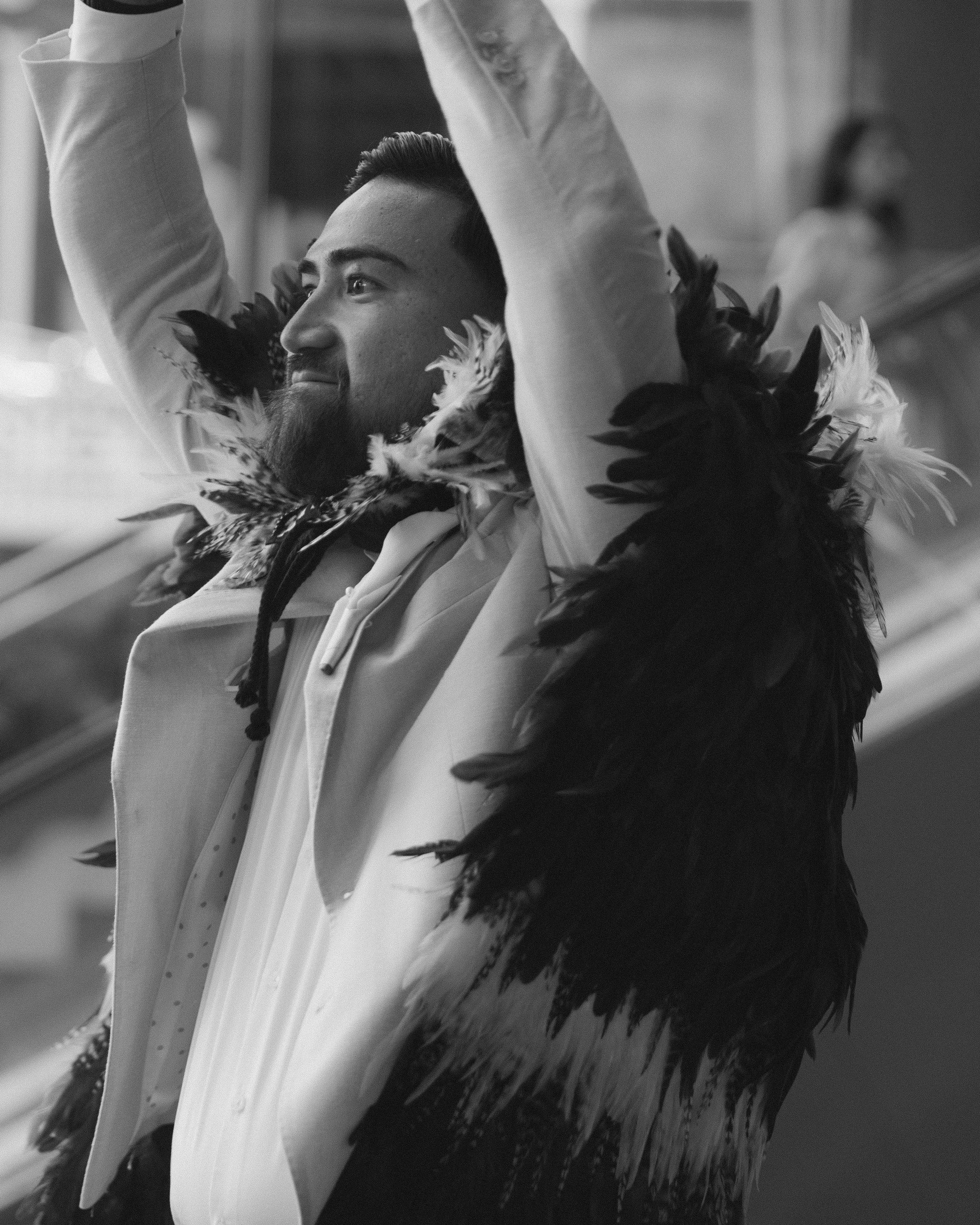 A man with a beard and mustache wearing a feathered necklace, raising his arms, in a celebratory moment.