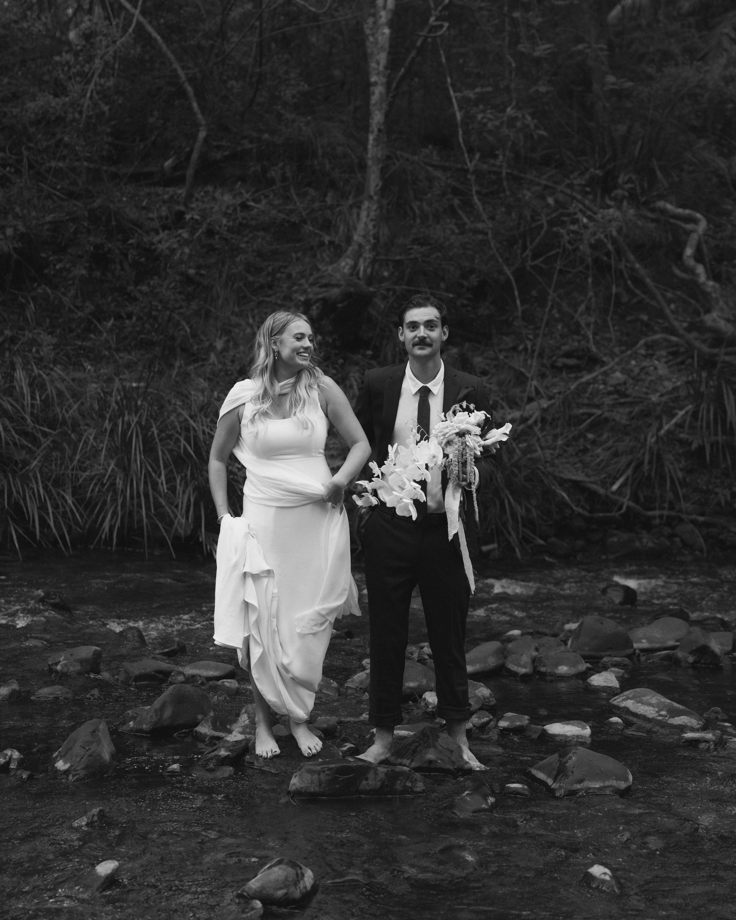 A black and white photo of a bride and groom standing barefoot in a shallow rocky stream, surrounded by trees and foliage, holding hands and smiling.