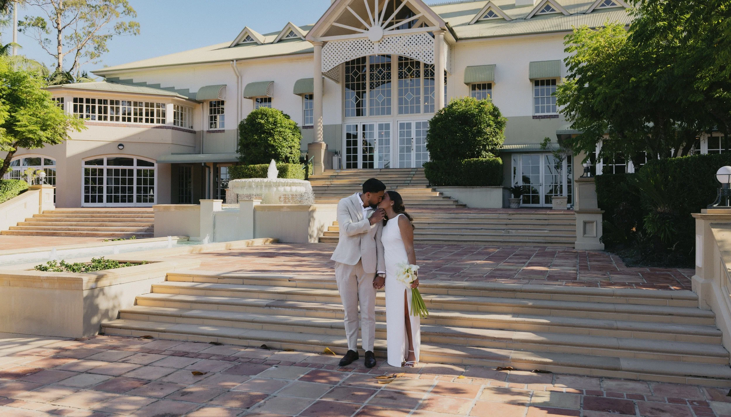 A couple dressed in wedding attire sharing a kiss outside a large, elegant white mansion with steps, a fountain, and well-maintained landscaping.