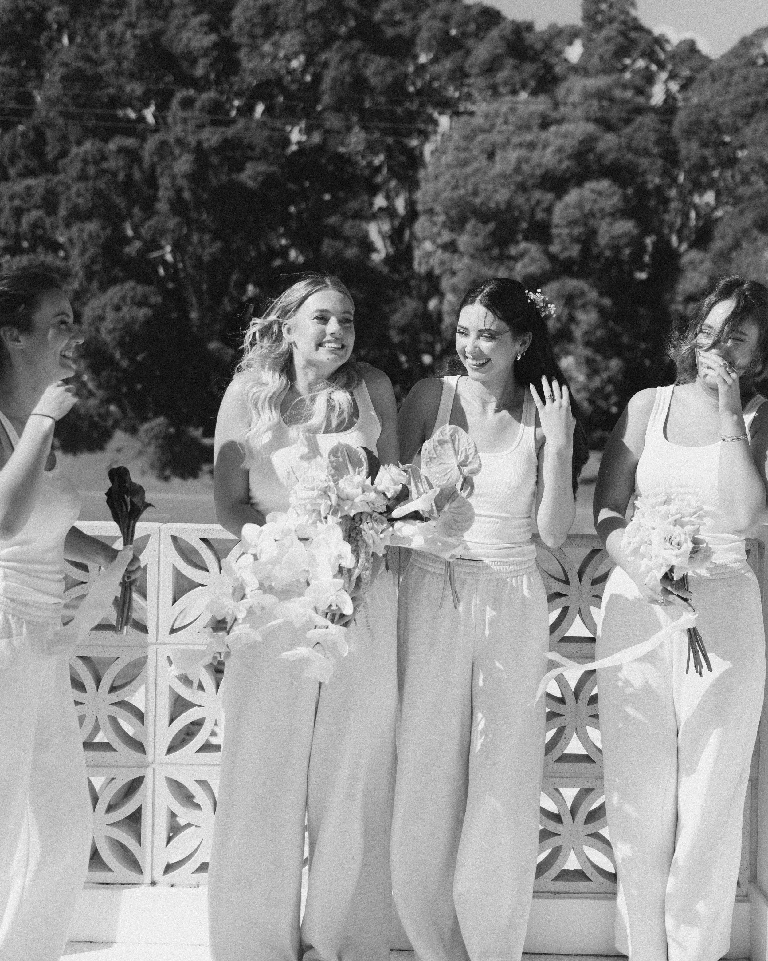 Four women in athletic wear, smiling and laughing, standing outdoors against a decorative fence with trees in the background; one woman holds a bouquet of flowers.