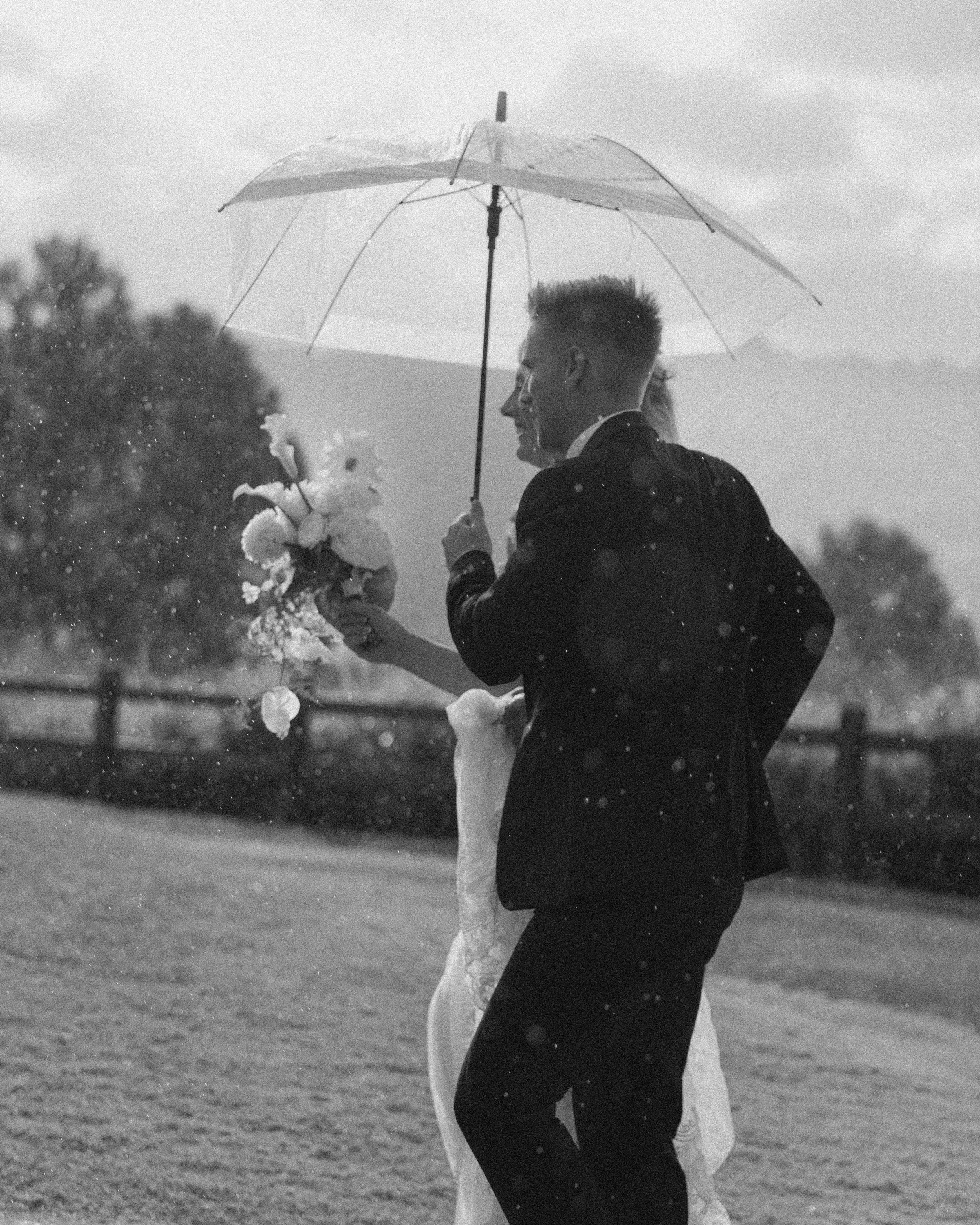 A black and white photo of a bride and groom dancing outside in the rain, with the groom holding an umbrella and the bride holding a bouquet.