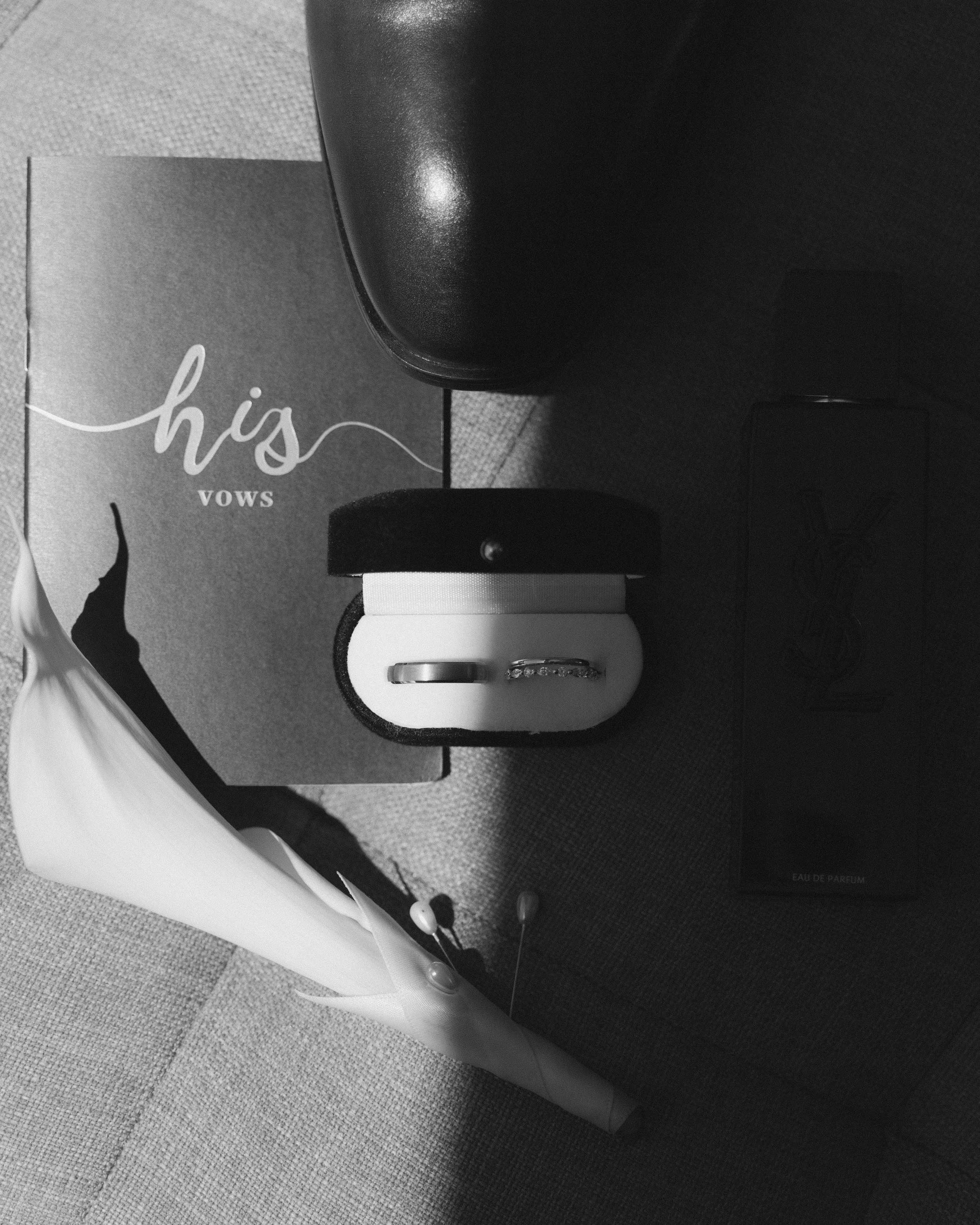 A black and white photo of a box of wedding rings, a 'his vows' card, a bouquet, a perfume bottle, and a pair of wedding shoes.