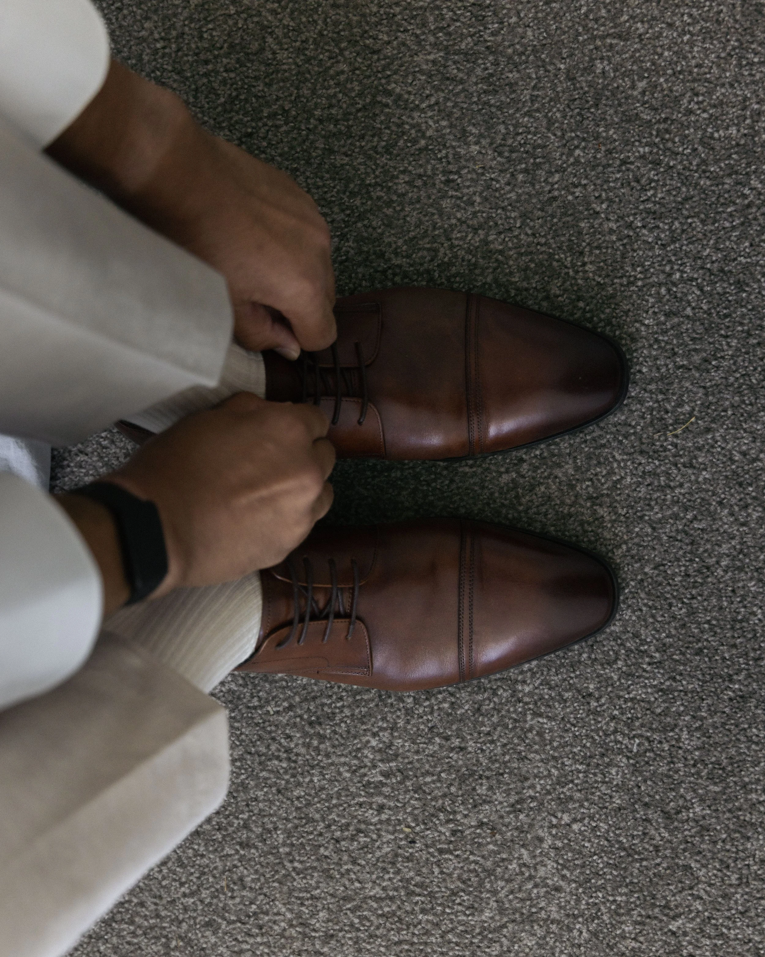 A person wearing tan trousers and cream-colored socks, tying their brown leather dress shoes on a gray carpeted floor.
