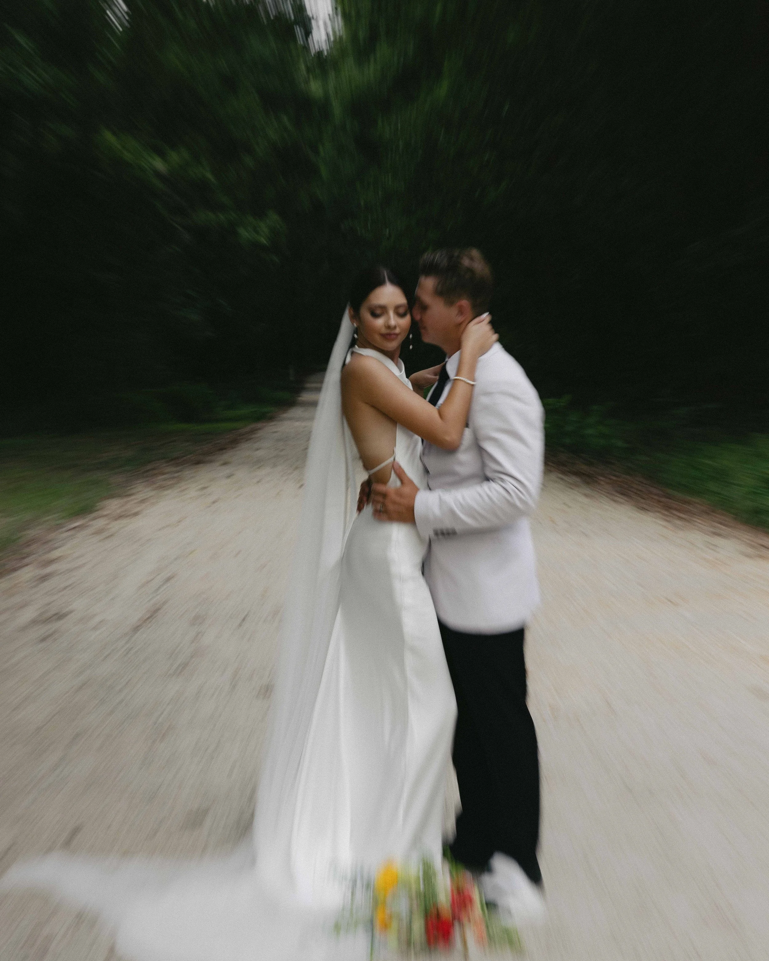 A bride and groom embrace on a sandy path surrounded by green foliage, with a blurred background creating a zoom effect