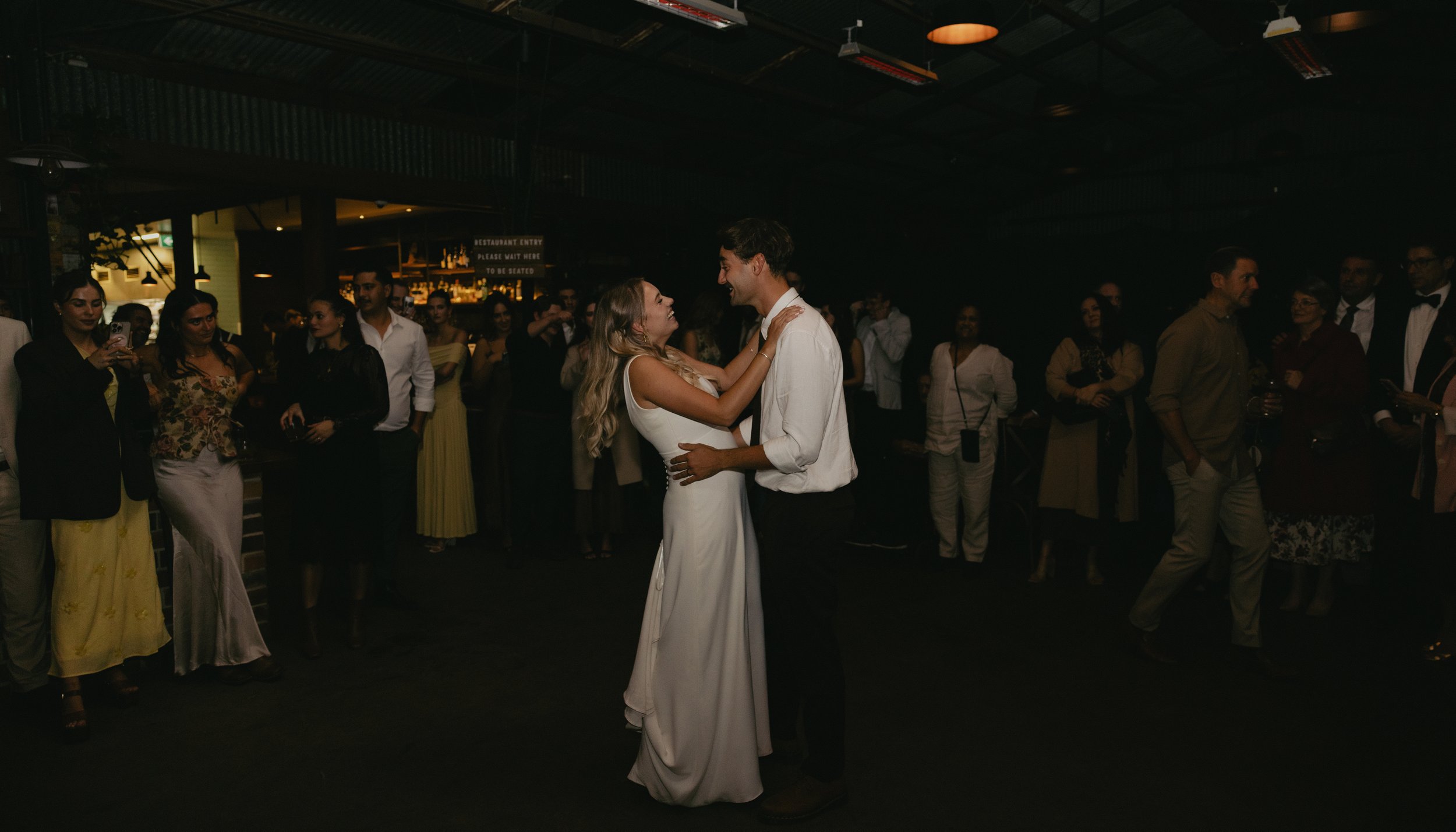 A bride and groom dance together at their wedding reception, surrounded by friends and family in a dimly lit venue.