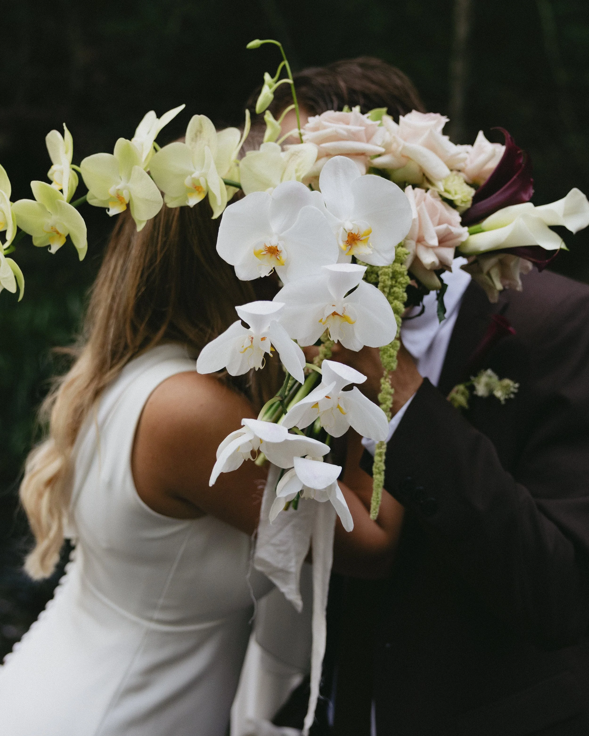 A couple kissing behind a large bouquet of white orchids and other flowers during a wedding.