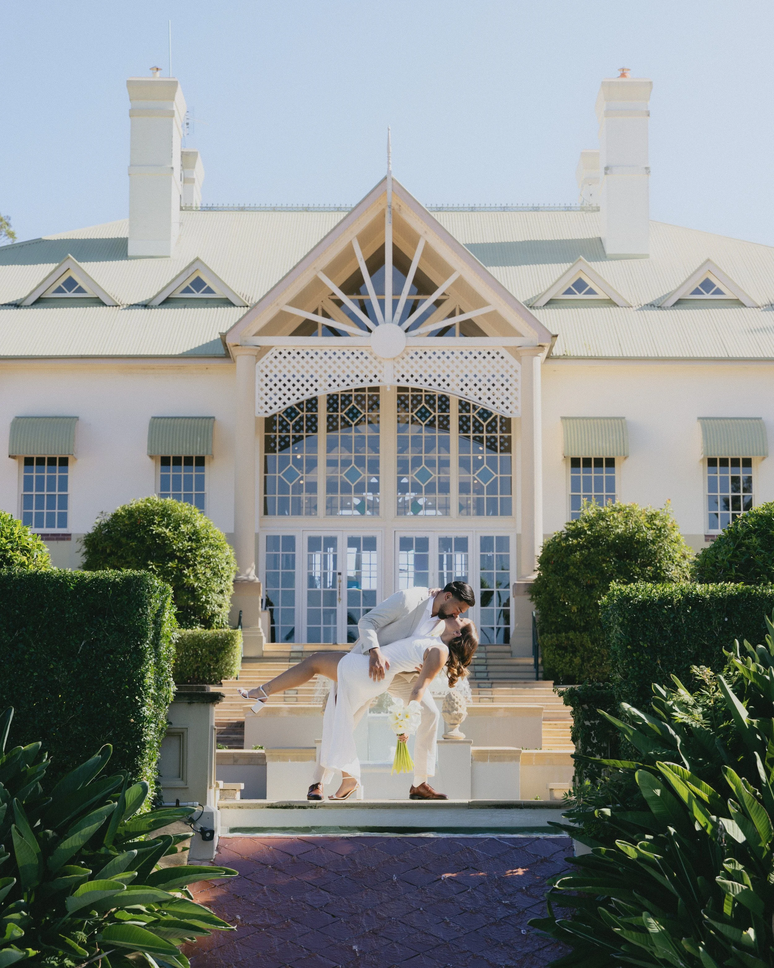 A couple sexually entwined on the steps of a large white house with elaborate architecture, including peaked rooflines, large windows, and surrounding lush greenery.