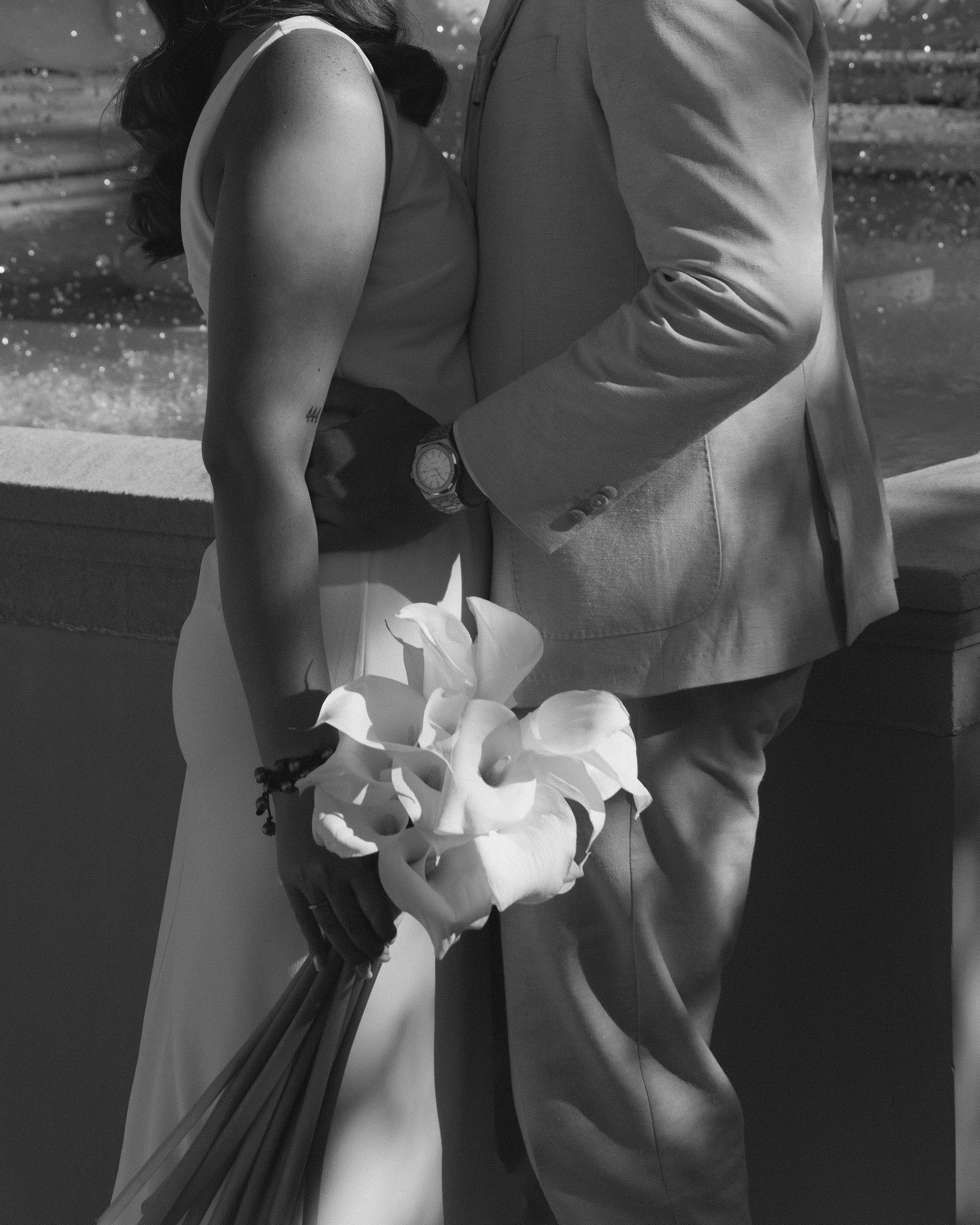 A couple holding hands, with the woman holding a bouquet of flowers, standing by a water feature, in black and white.