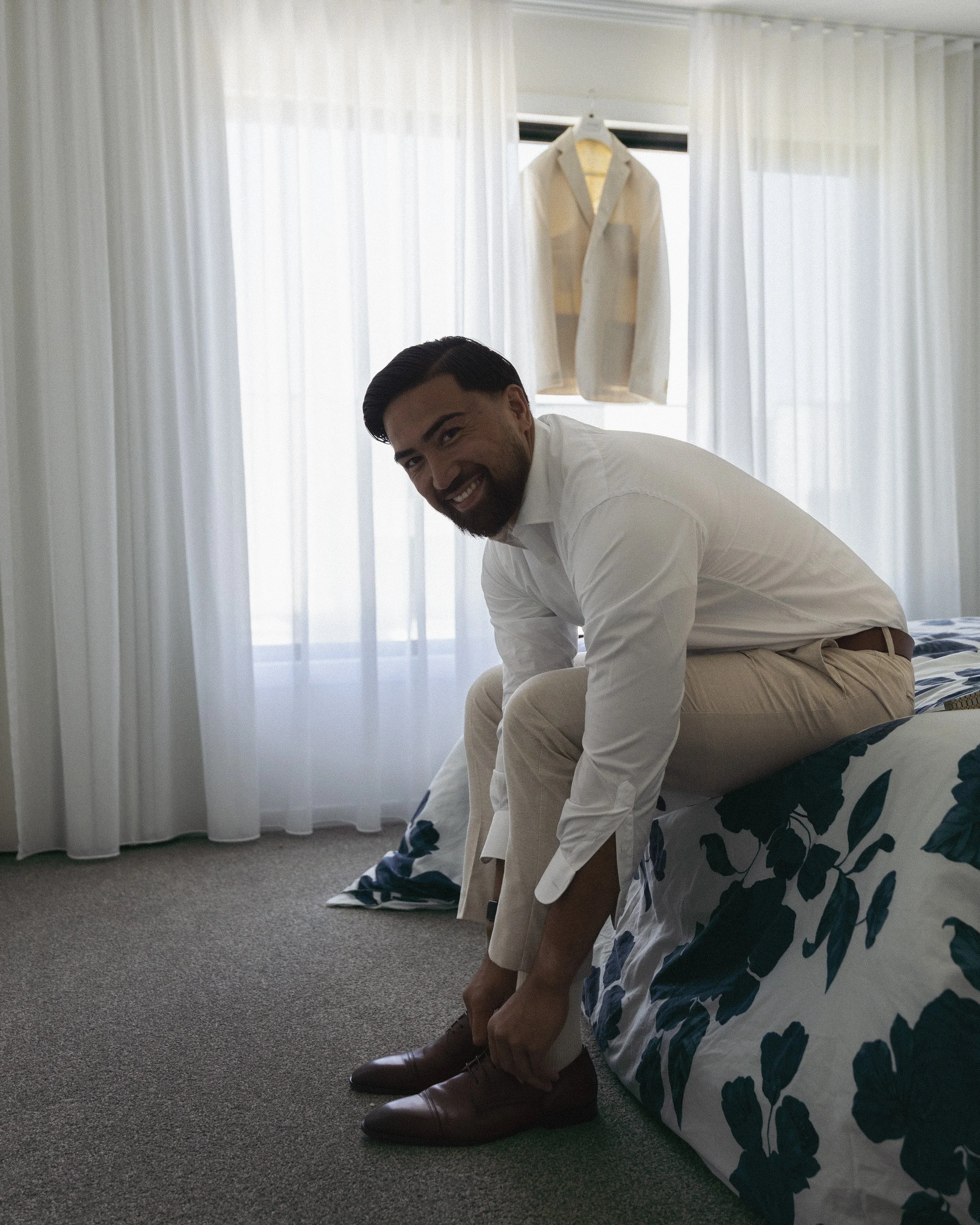 A man in a white shirt and beige pants sitting on a bed, smiling while tying his brown shoes in a bright room with white curtains and an open window.