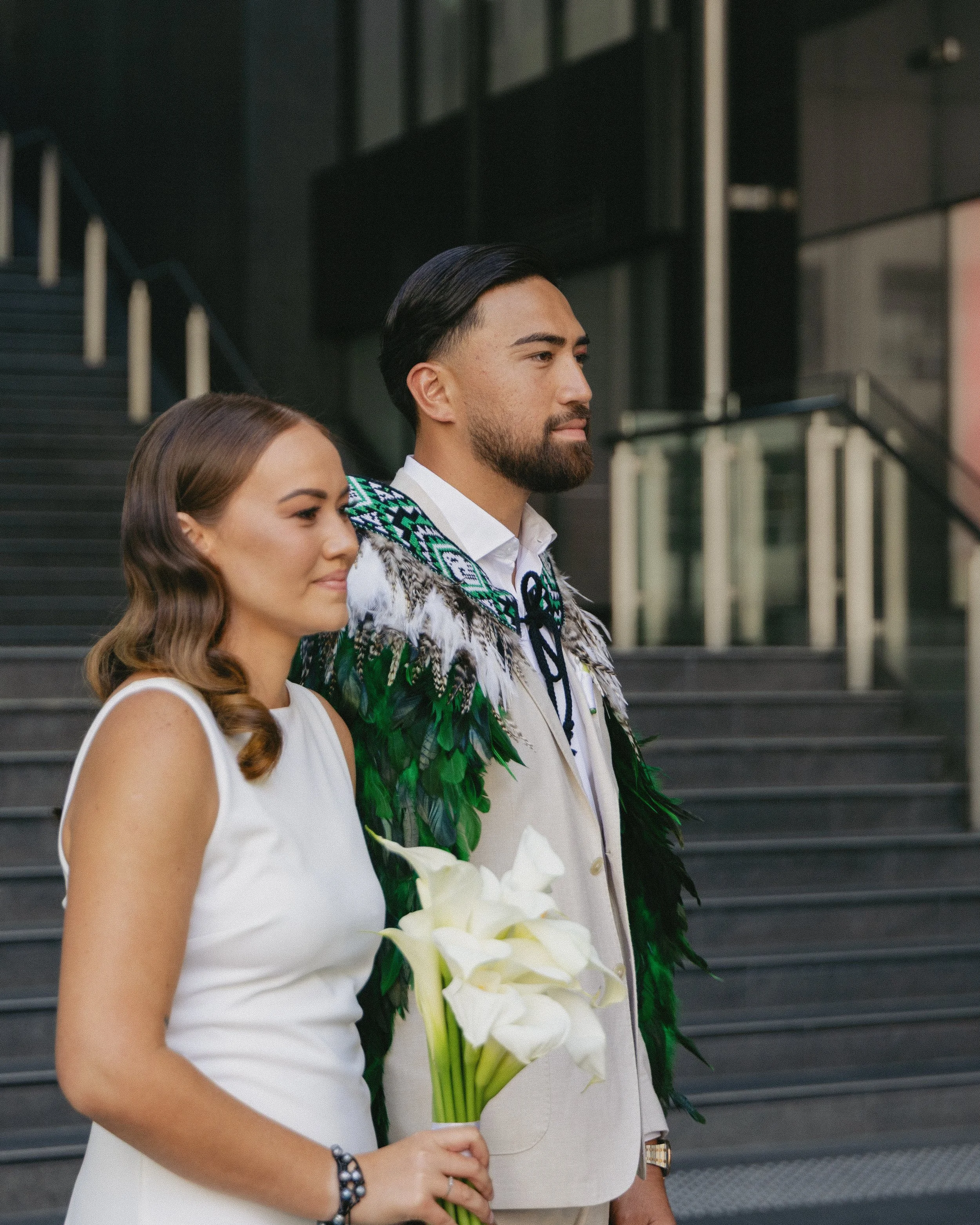 A couple standing together outdoors, with the woman holding a bouquet of white calla lilies. The man is wearing traditional Indigenous attire with feather decorations. They are positioned in front of a modern building with stairs and glass railing.