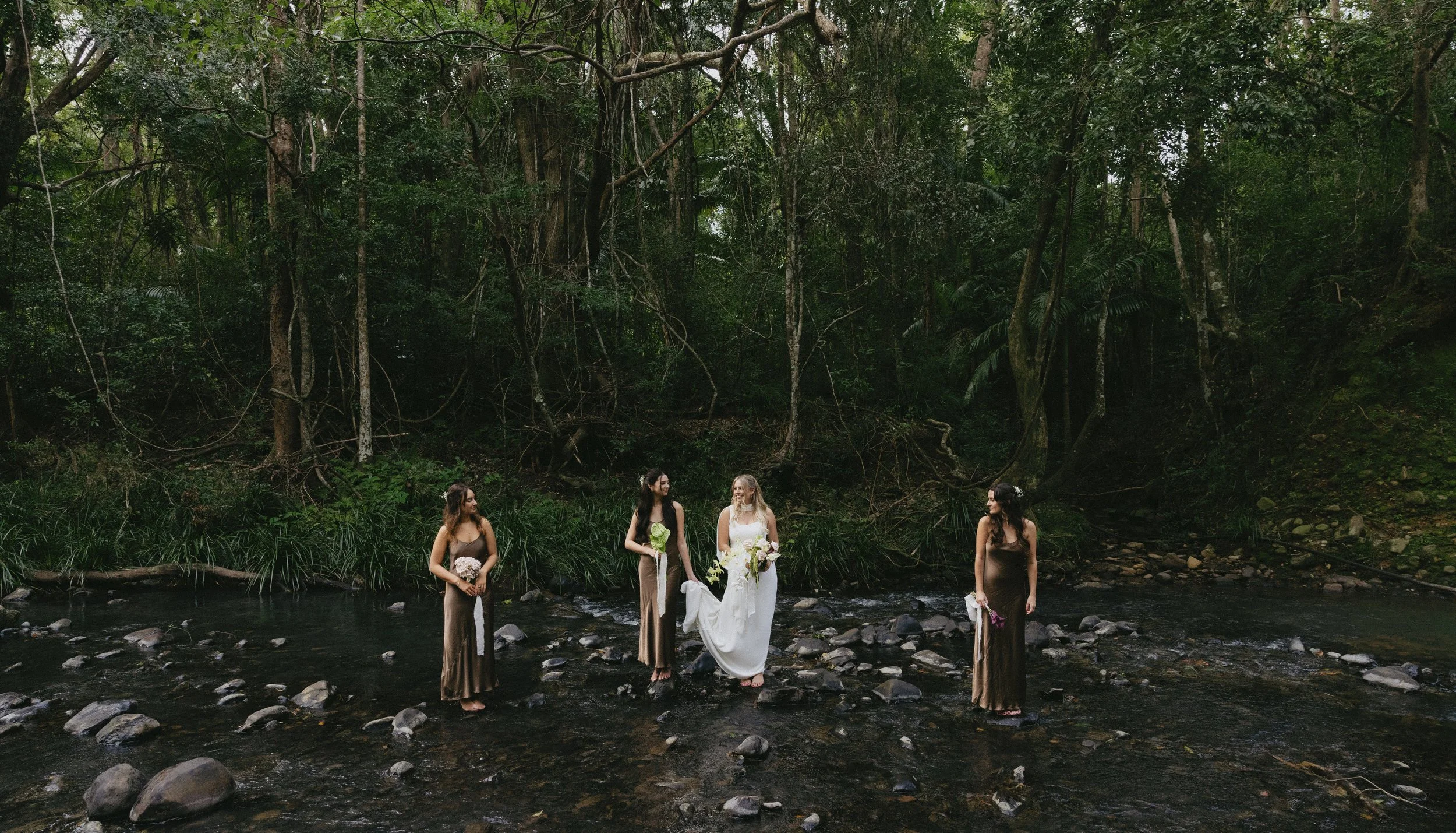 A wedding ceremony taking place in a forest by a river, with four women dressed in formal attire and holding flowers, surrounded by tall trees and lush greenery.