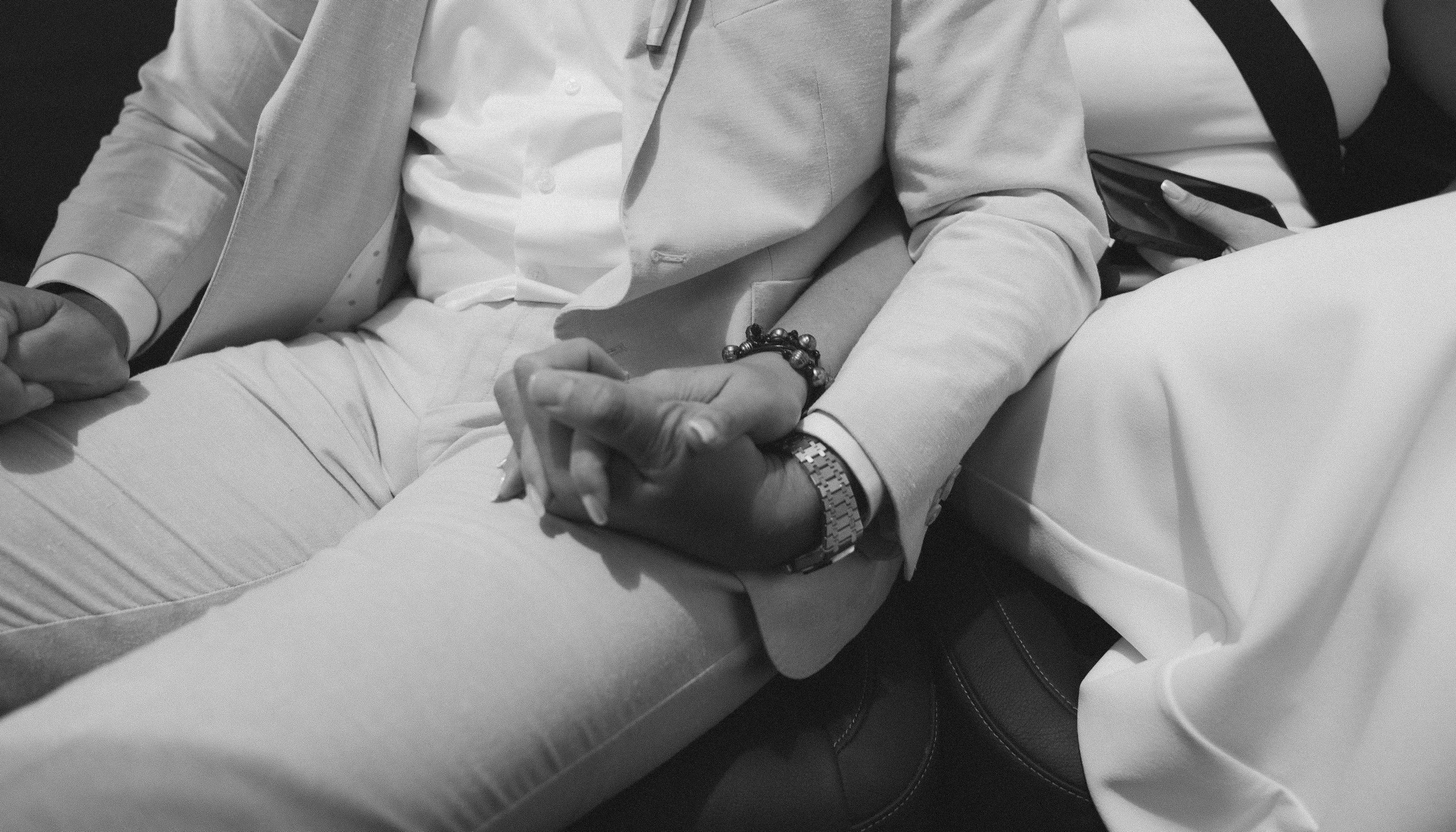 Close-up of two people sitting next to each other, holding hands on their laps, one wearing a light-colored blazer and the other in white clothing, with visible jewelry and a smartphone.