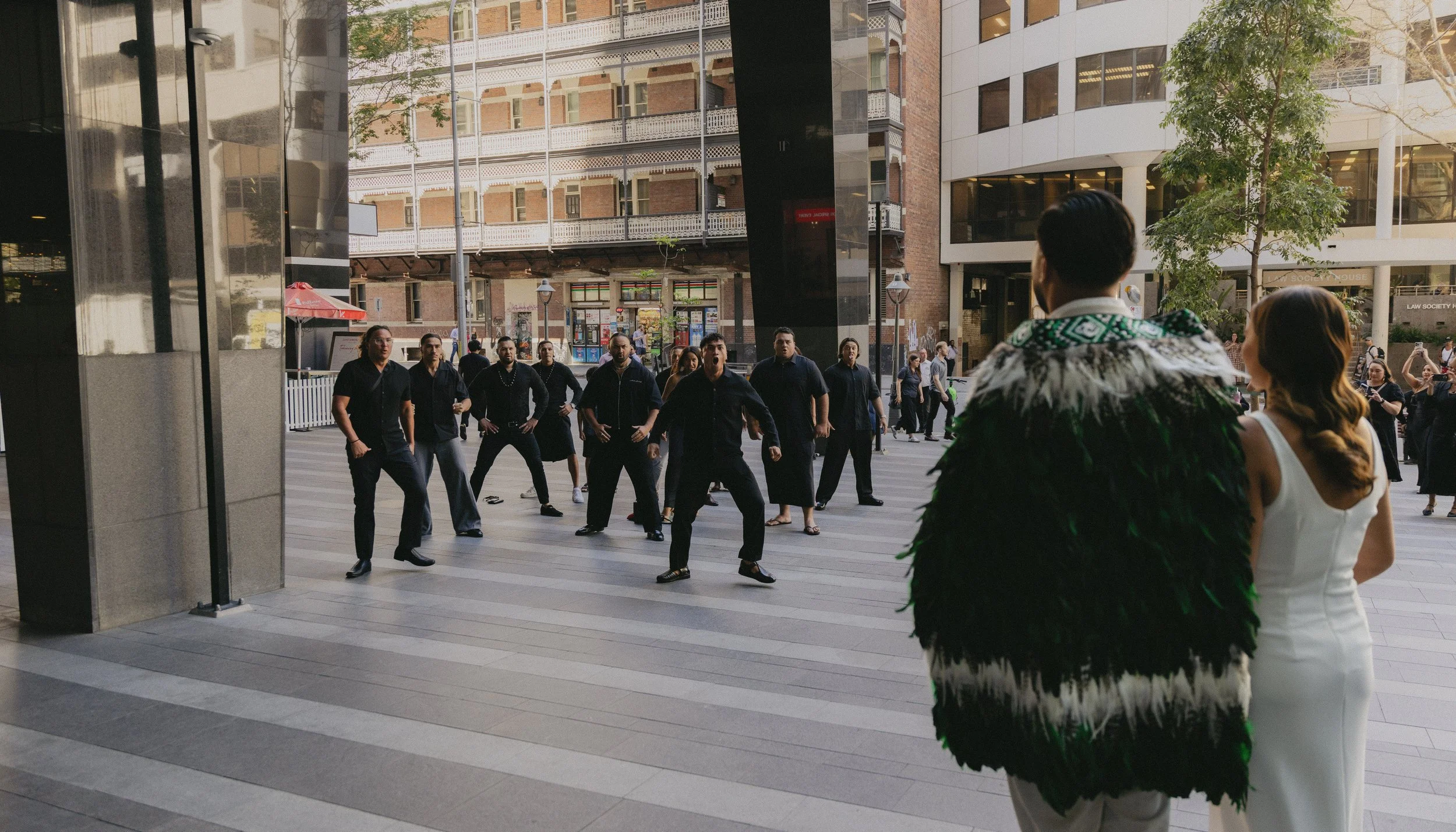 Couple watching a dance performance on a city street, with dancers in black outfits performing in front of tall buildings and a group of onlookers taking photos in the background.