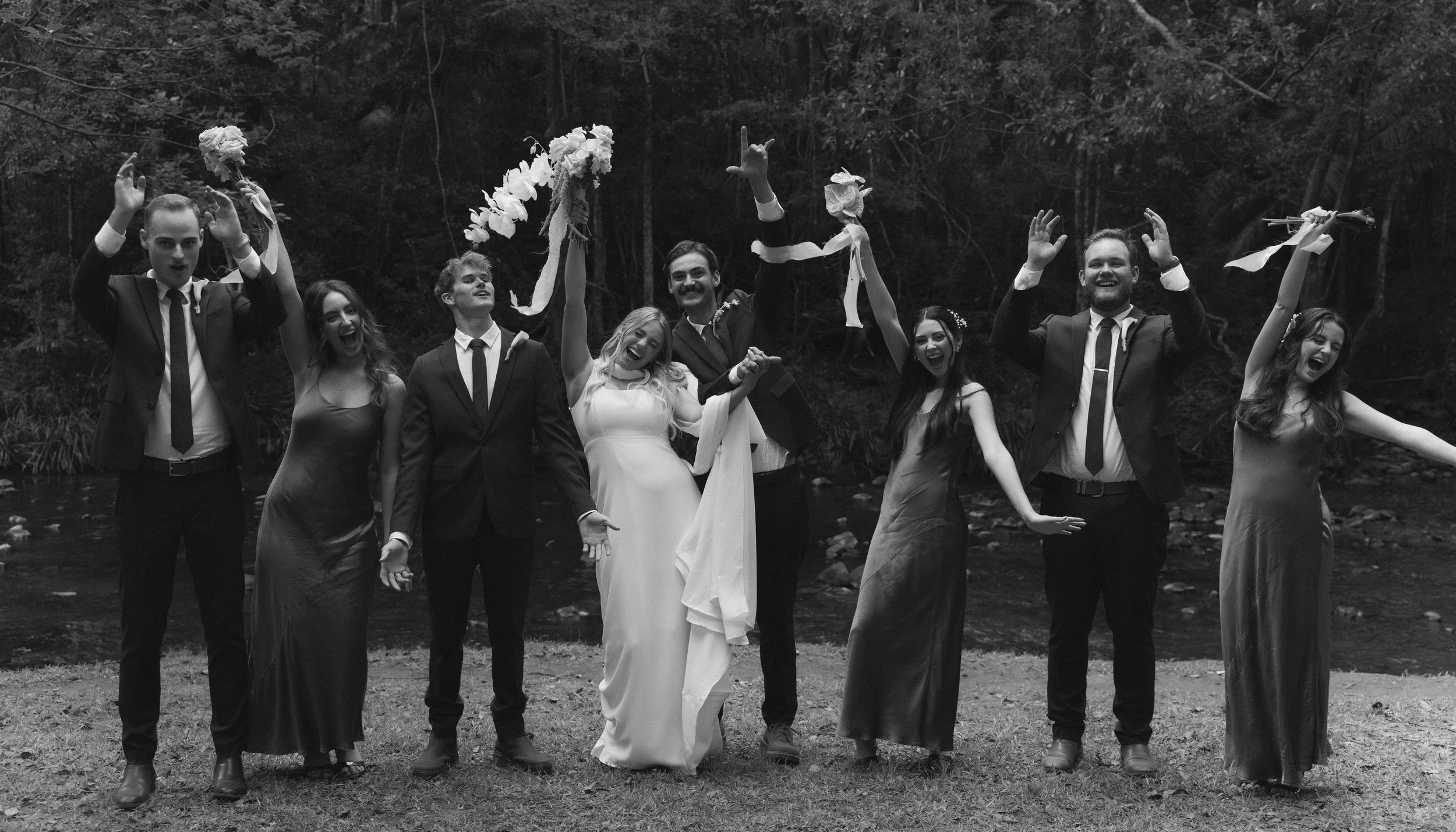 Group of people celebrating outdoors near a river, dressed in formal attire, with some holding floral decorations.
